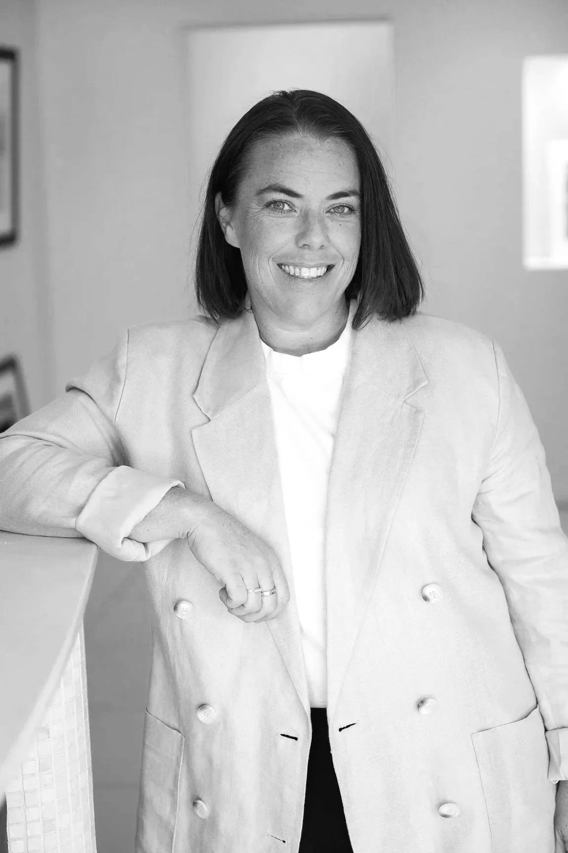Black and white photo of Carrie McInerney,  CEO of Horner Management, smiling, wearing a double-breasted blazer and a white top, leaning on a table with her right arm.