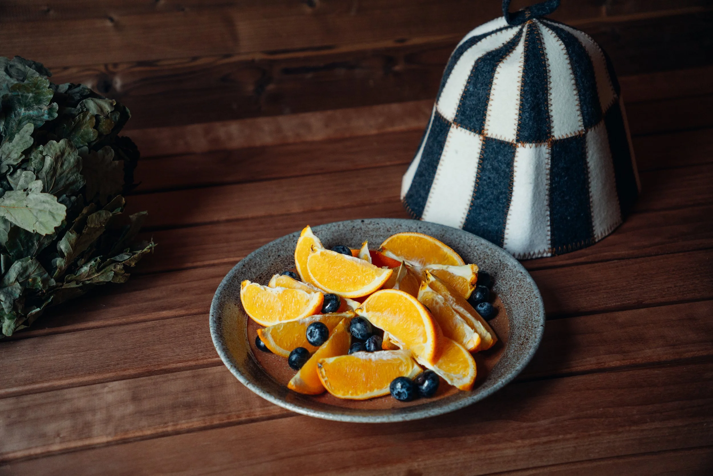 A plate of orange slices and blueberries on a wooden table, with a plaid cloth cap nearby and a bunch of leafy greens to the side.