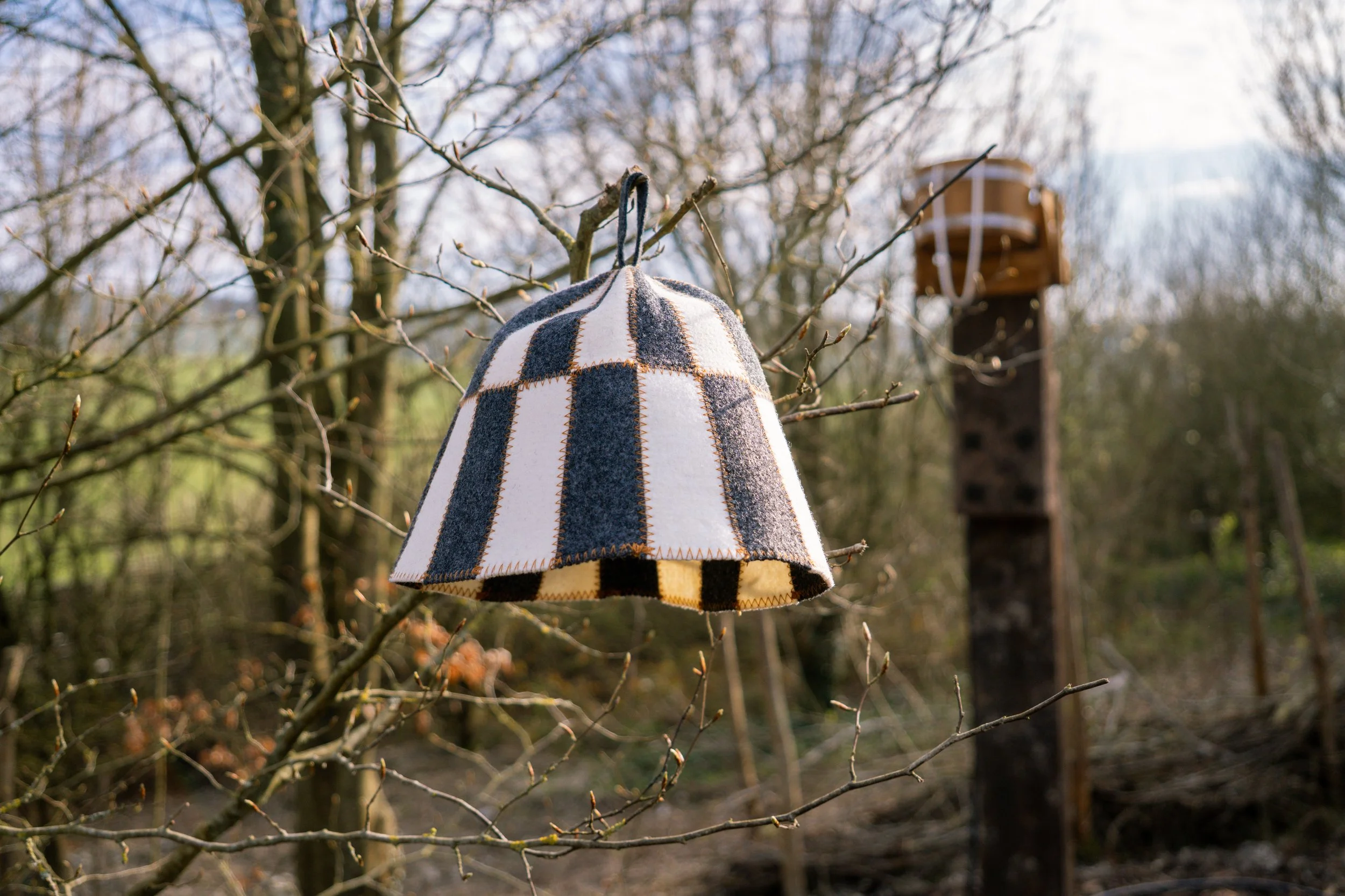 A lampshade made of checkered fabric hanging from a tree branch outdoors in a wooded area with a wooden birdhouse in the background.