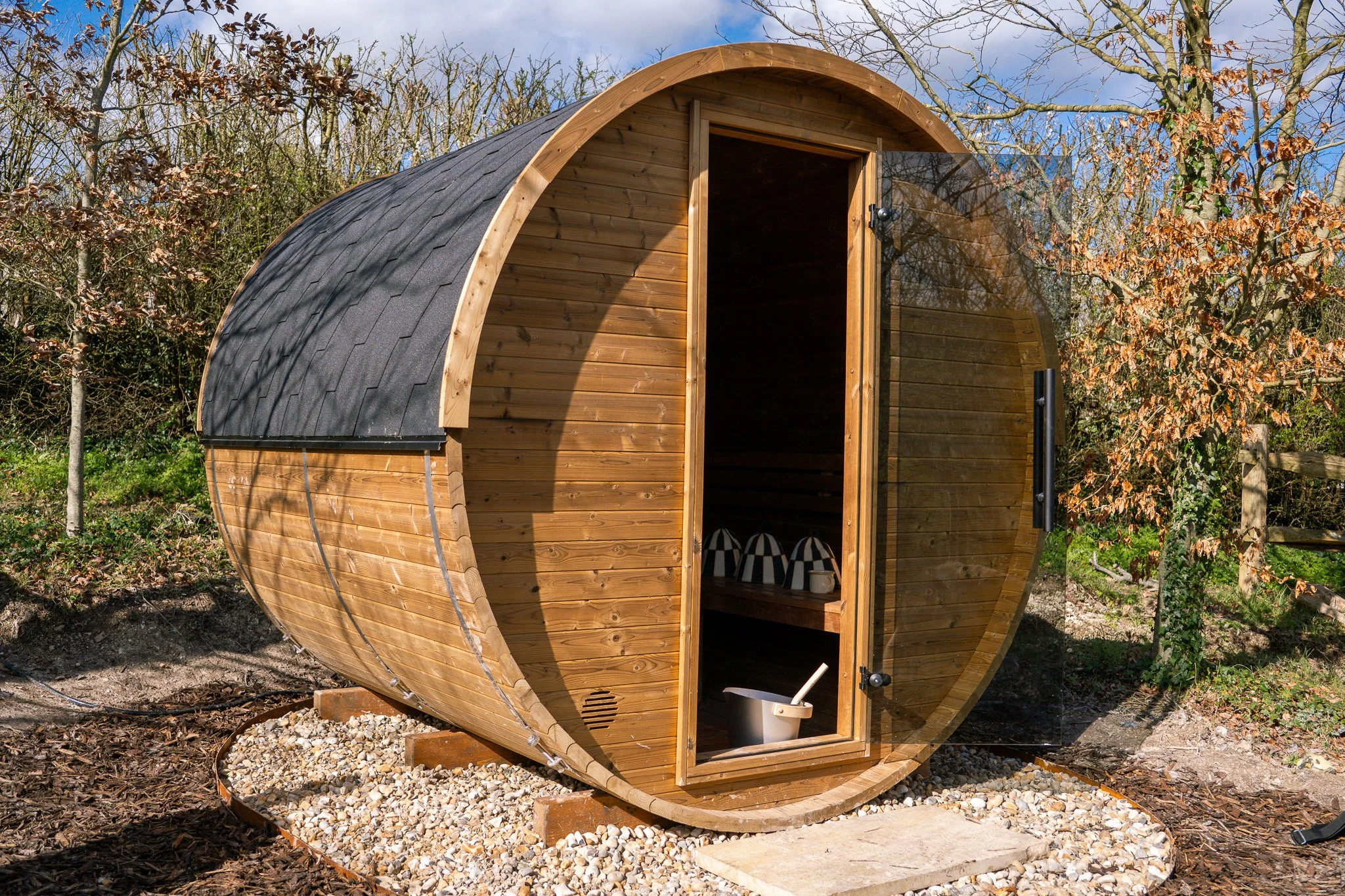 A small, rounded wooden outdoor sauna on a gravel base in a garden, with a door open revealing interior with sauna stones and a bucket with a dipper.