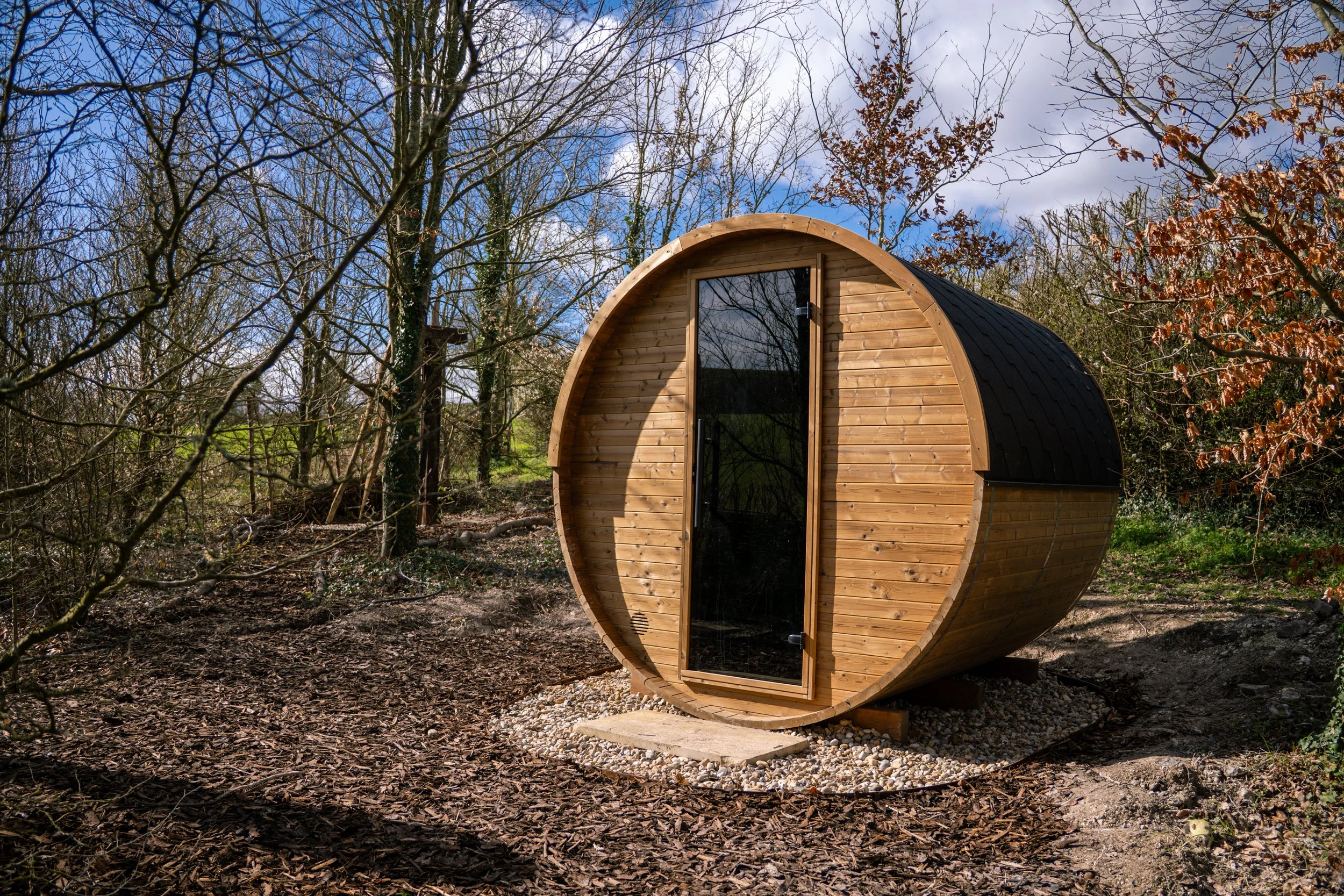 A small, round wooden cabin with a black shingled roof and a glass door, set in a wooded area on a gravel foundation.