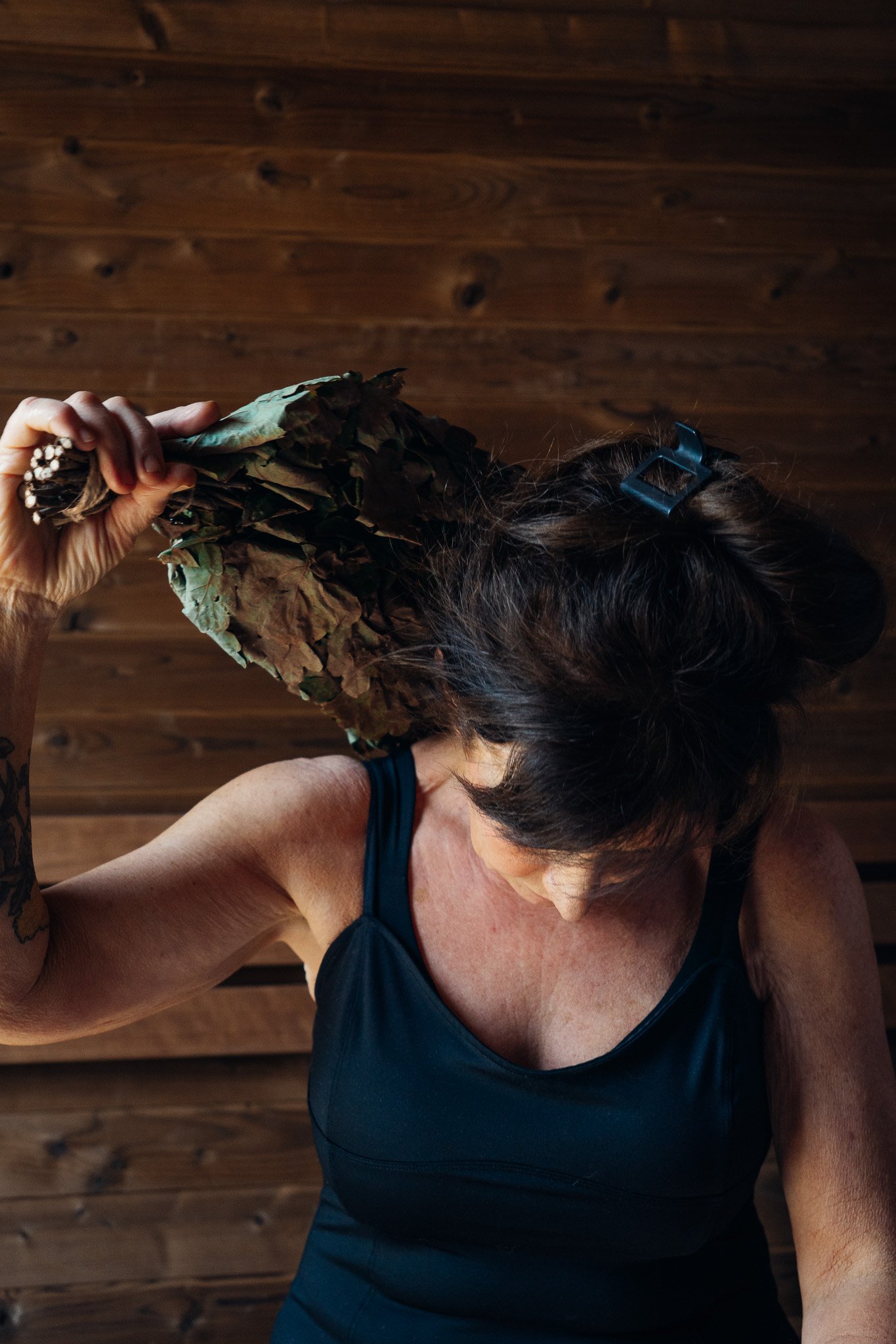 A woman holds a bundle of dried leaves or herbs over her shoulder, with her head slightly bowed, standing in front of a wooden wall.