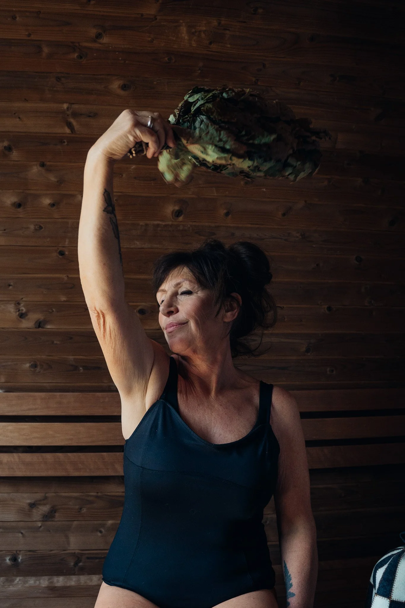 A woman in a black tank top raising her arm with a camouflage hat, standing against a wooden wall.
