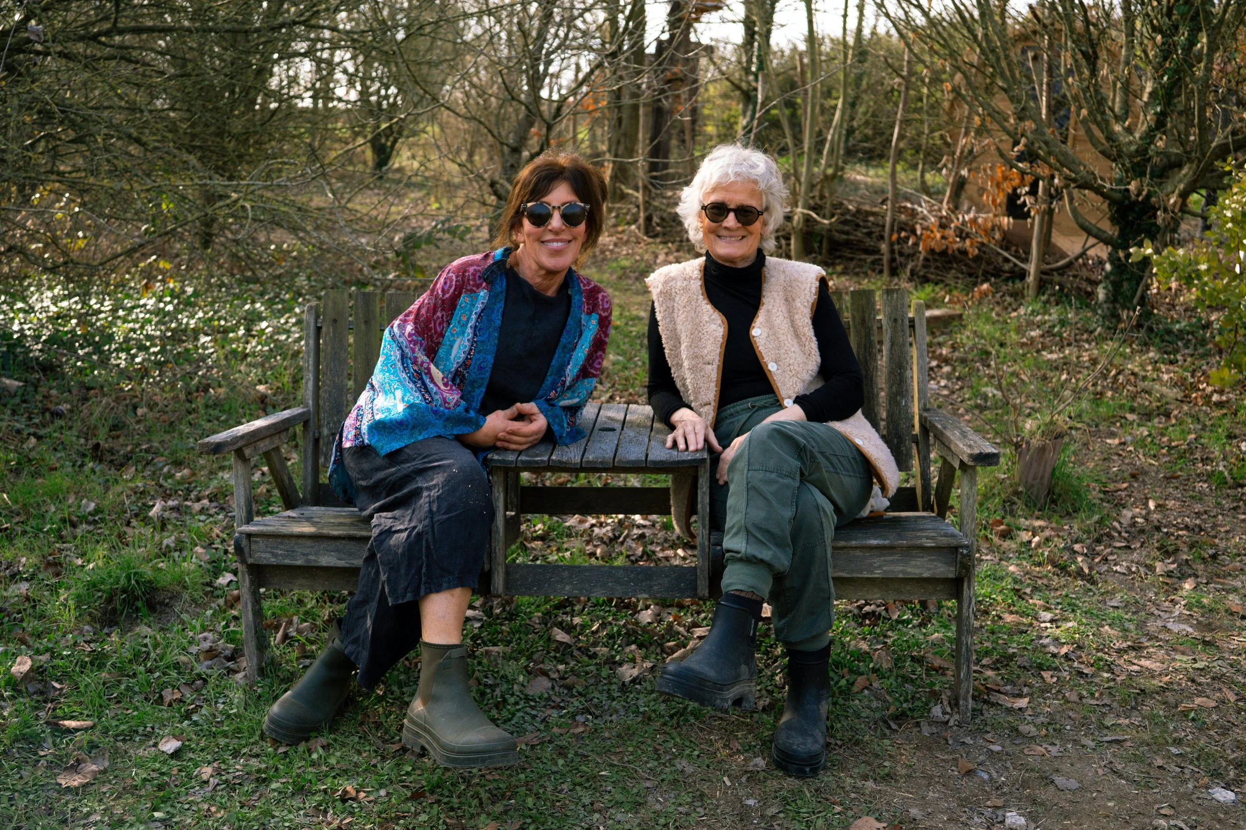 Two women sitting on a wooden bench outdoors, surrounded by trees and foliage, smiling at the camera.