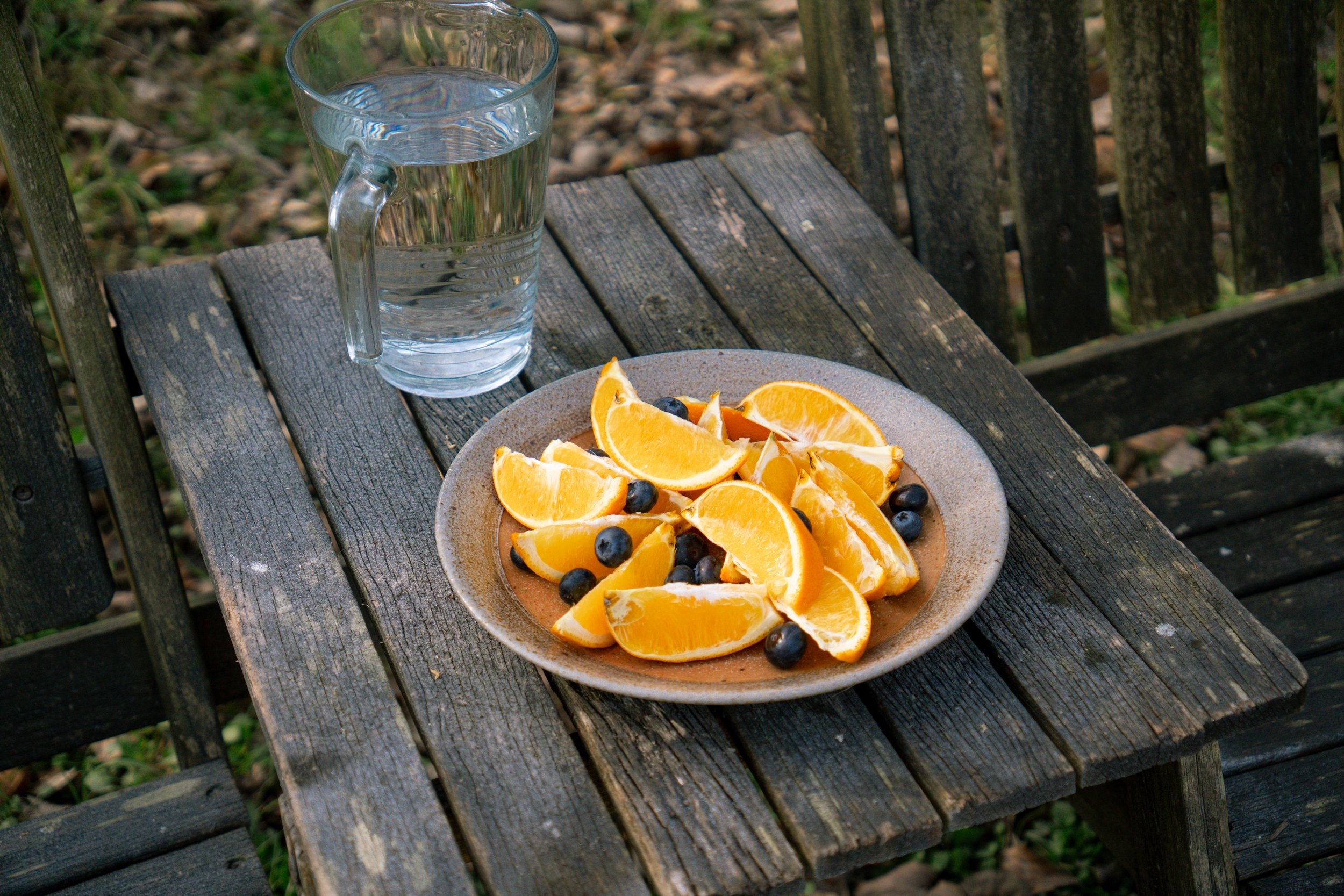 A rustic wooden outdoor table with a clear glass pitcher of water and a plate of orange slices and blueberries.