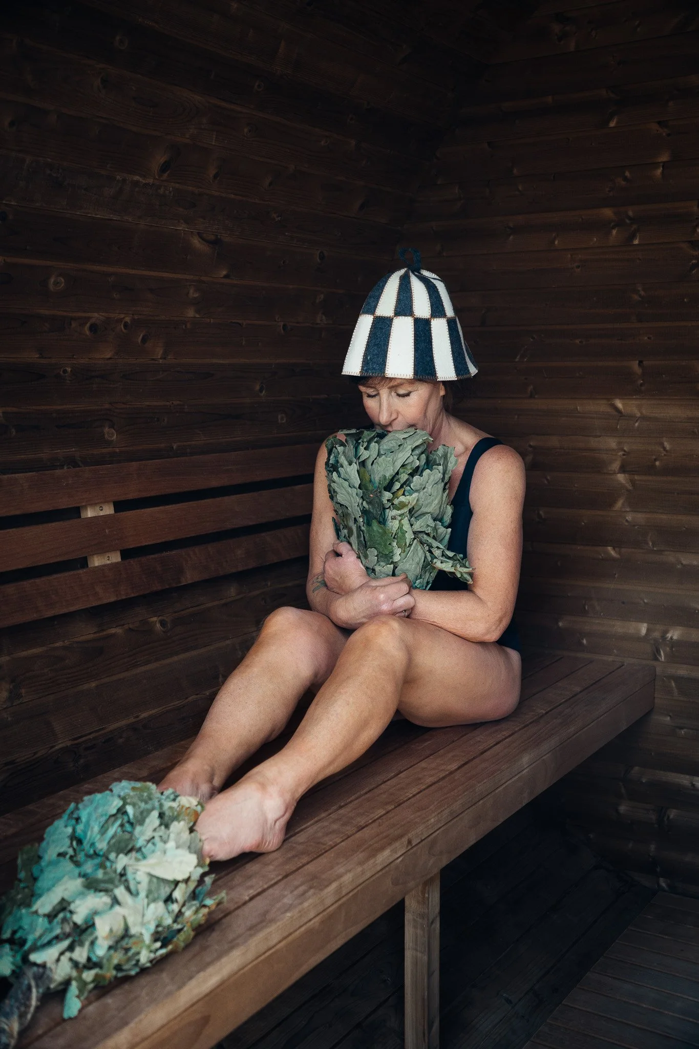 Woman sitting in a wooden sauna holding a bunch of leafy greens, wearing a blue and white striped bonnet, with more greens on the bench beside her.