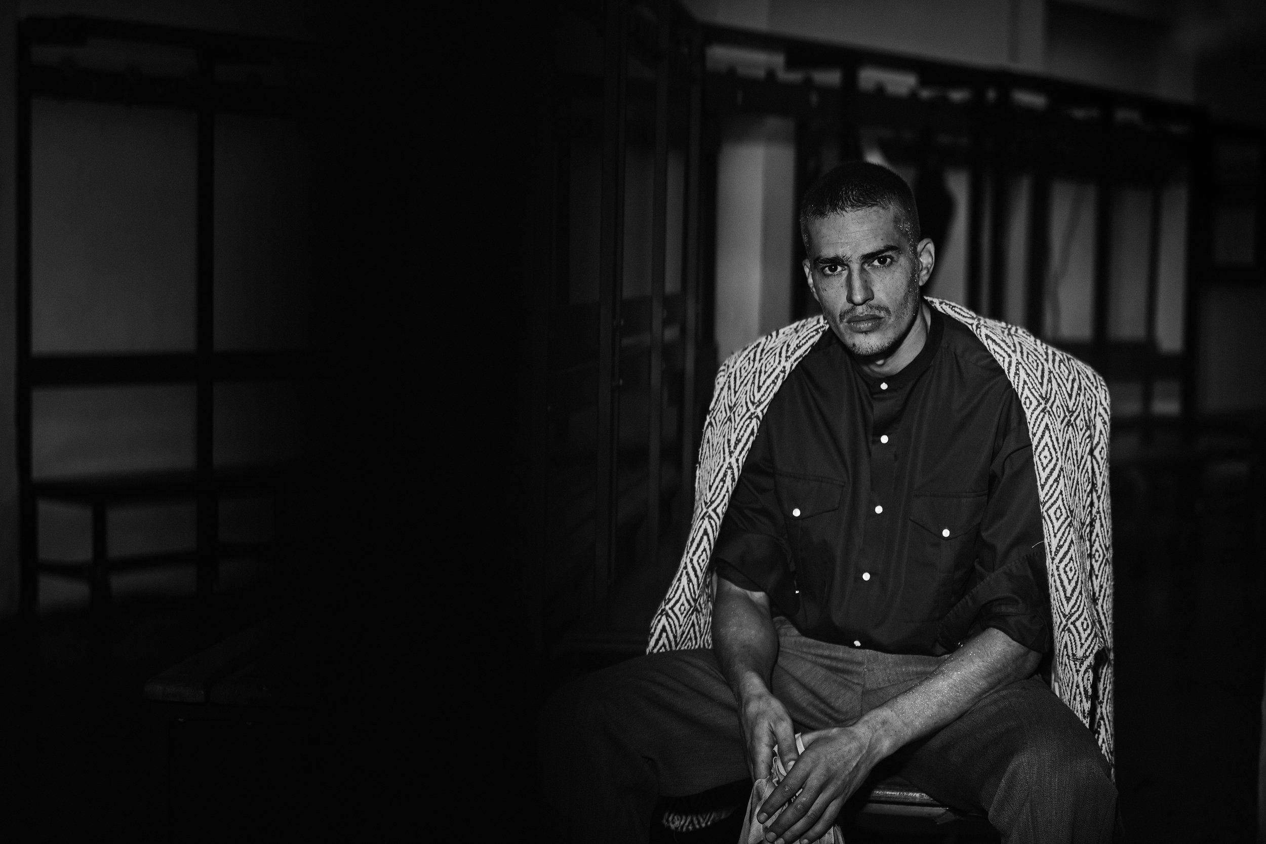 A black and white photo of a man sitting on the floor in a room, wearing a patterned jacket over a dark shirt, with a serious expression. The background contains metal shelving units.