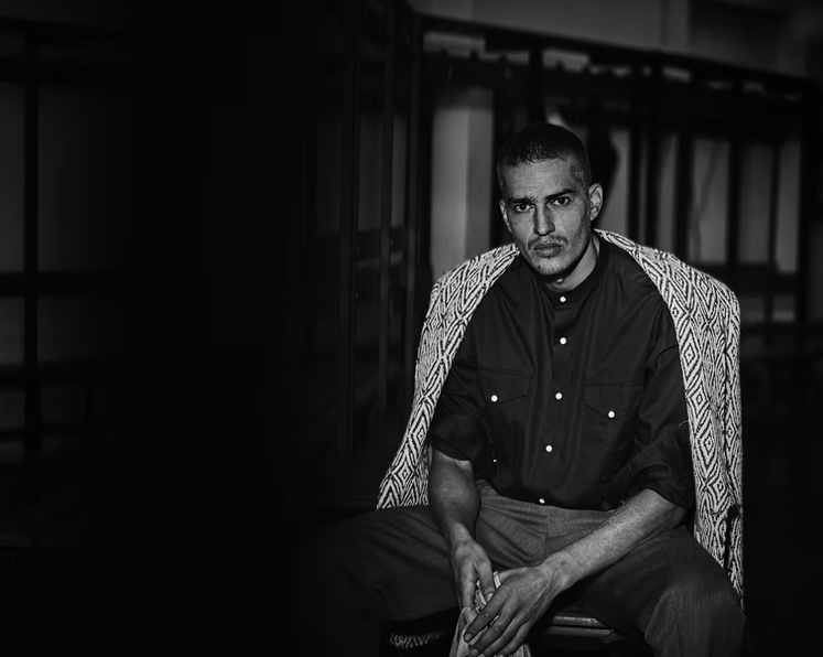 A black-and-white photo of a man sitting indoors, wearing a dark button-up shirt with rolled-up sleeves and a patterned jacket draped over his shoulders, looking directly at the camera.