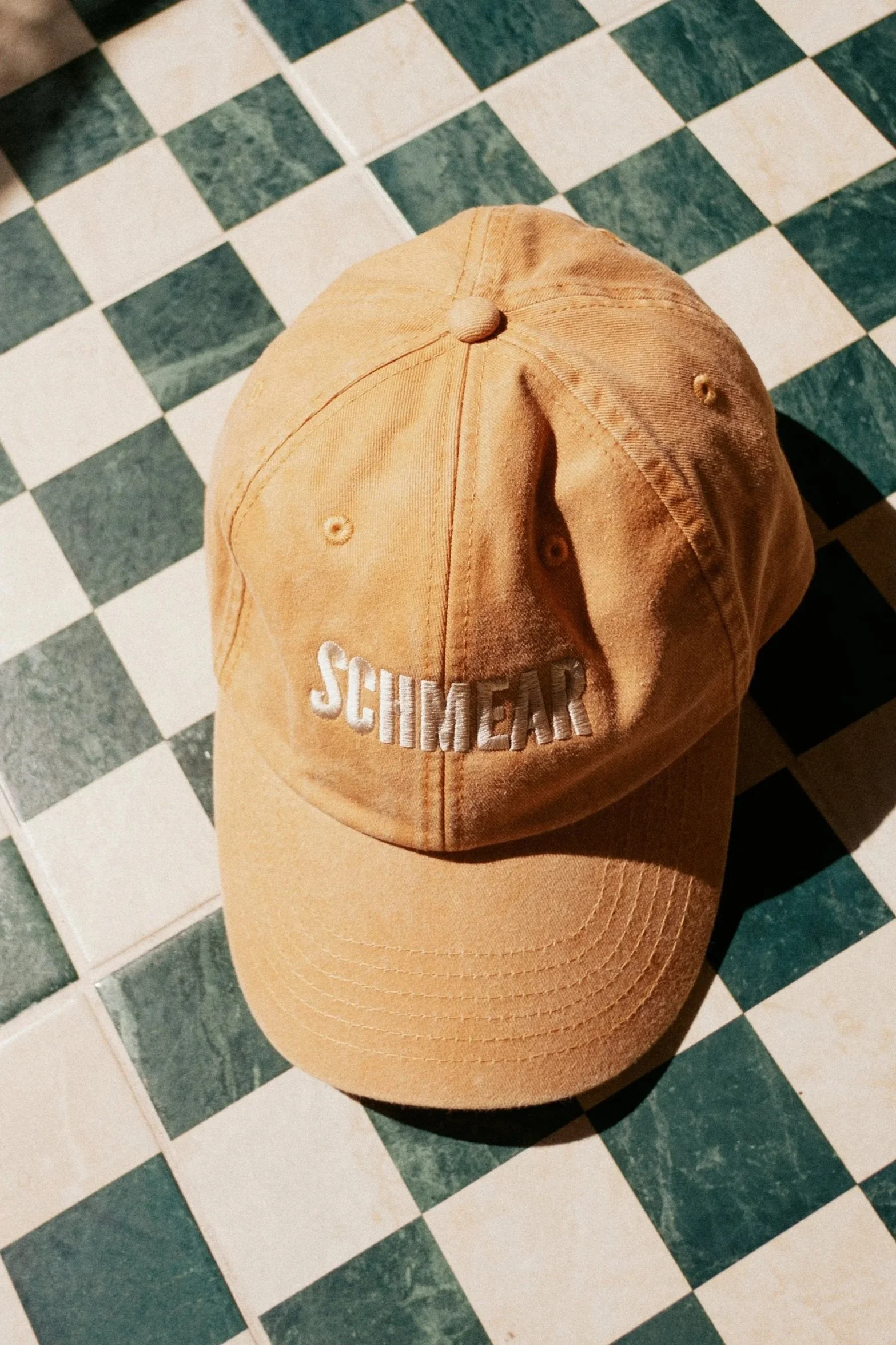 A tan baseball cap with the word "SCHMEAR" embroidered in white on the front, resting on a black and white checkered floor.