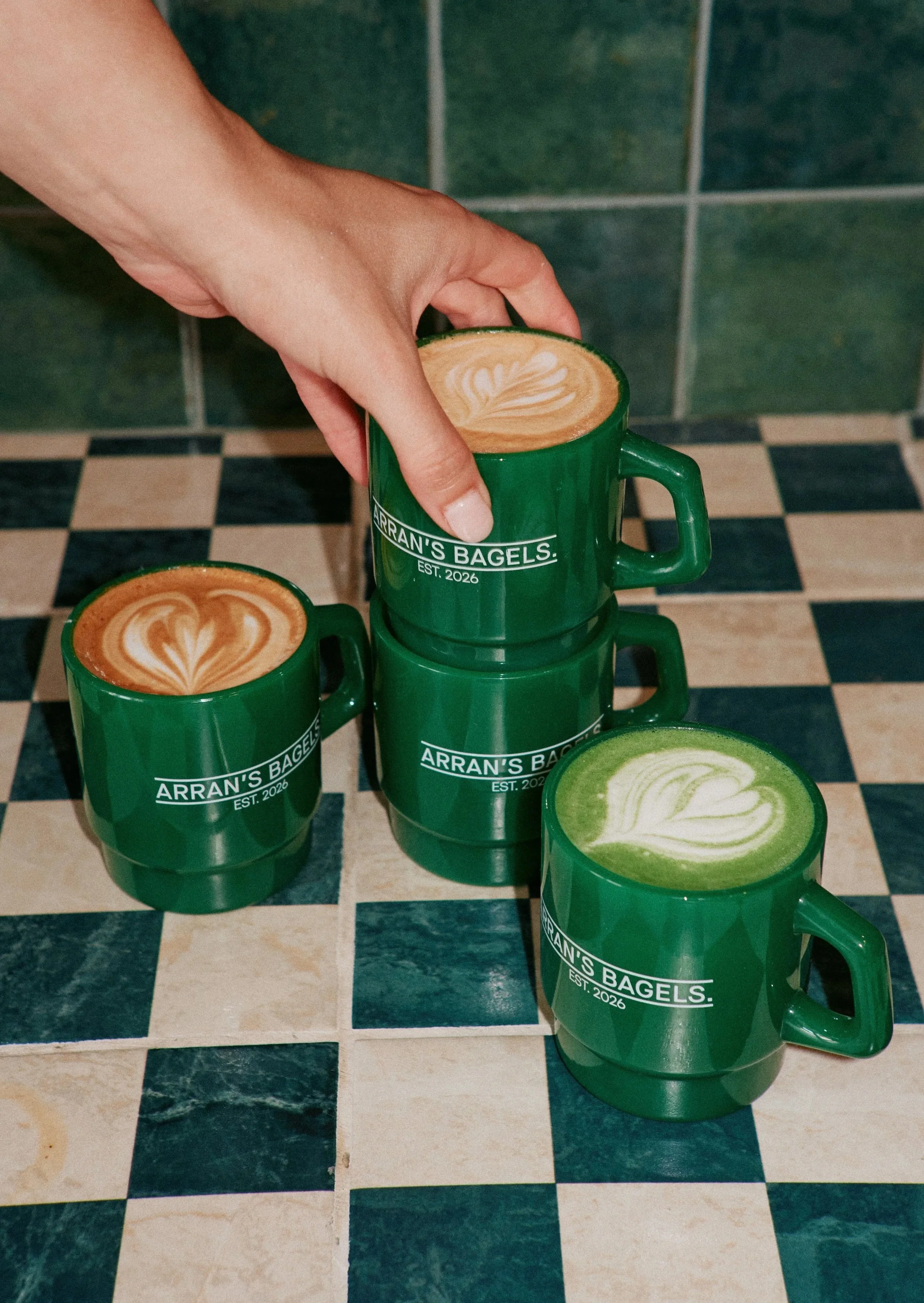 Multiple green coffee mugs with latte art, stacked on a black and white checkered tile floor, with a green tiled wall in the background, and the mugs display the text 'Arran's Bagels Est. 2026'.