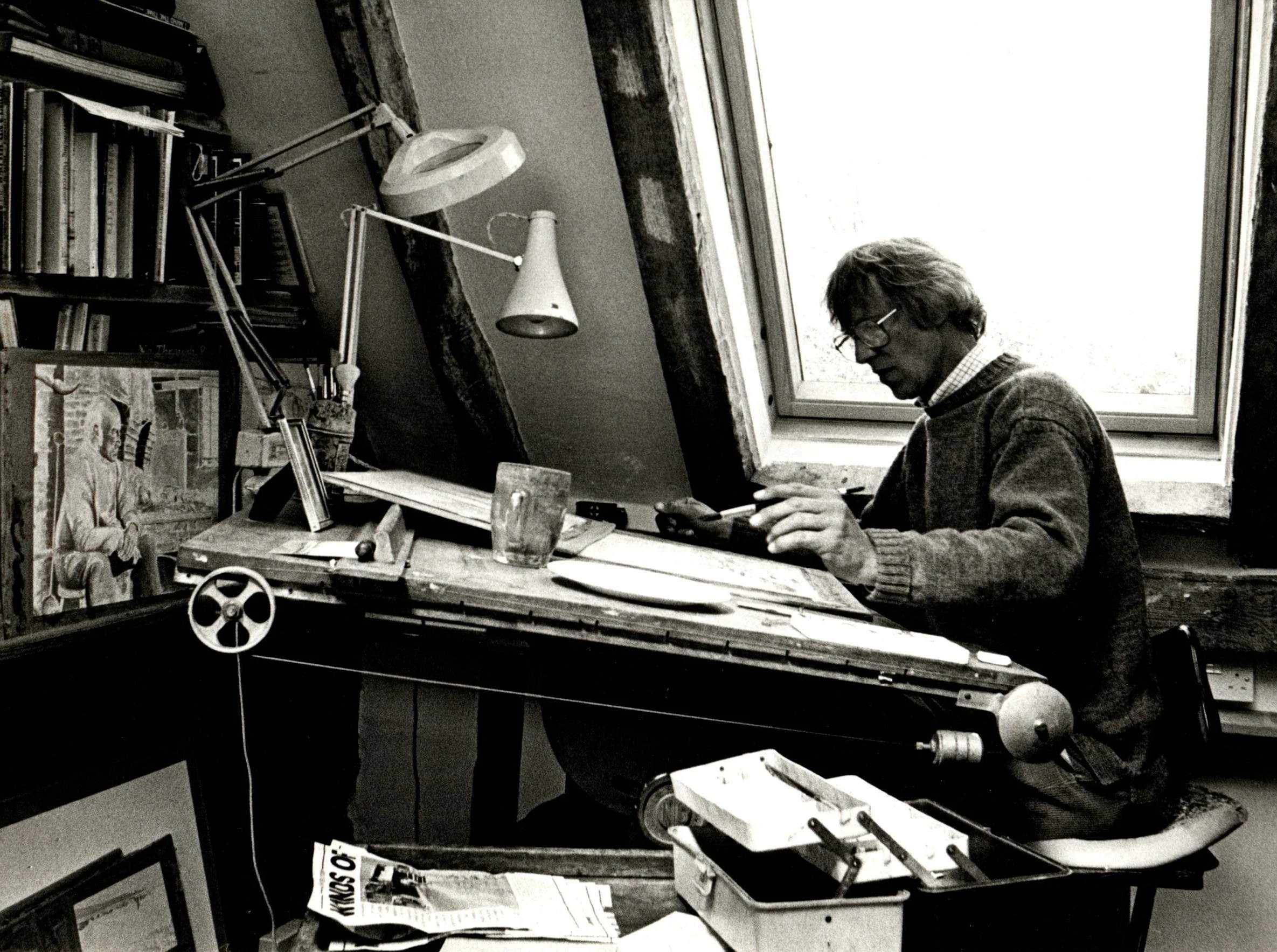 A man sitting at a cluttered desk in an attic room, looking at a smartphone, with a large window behind him letting in natural light.