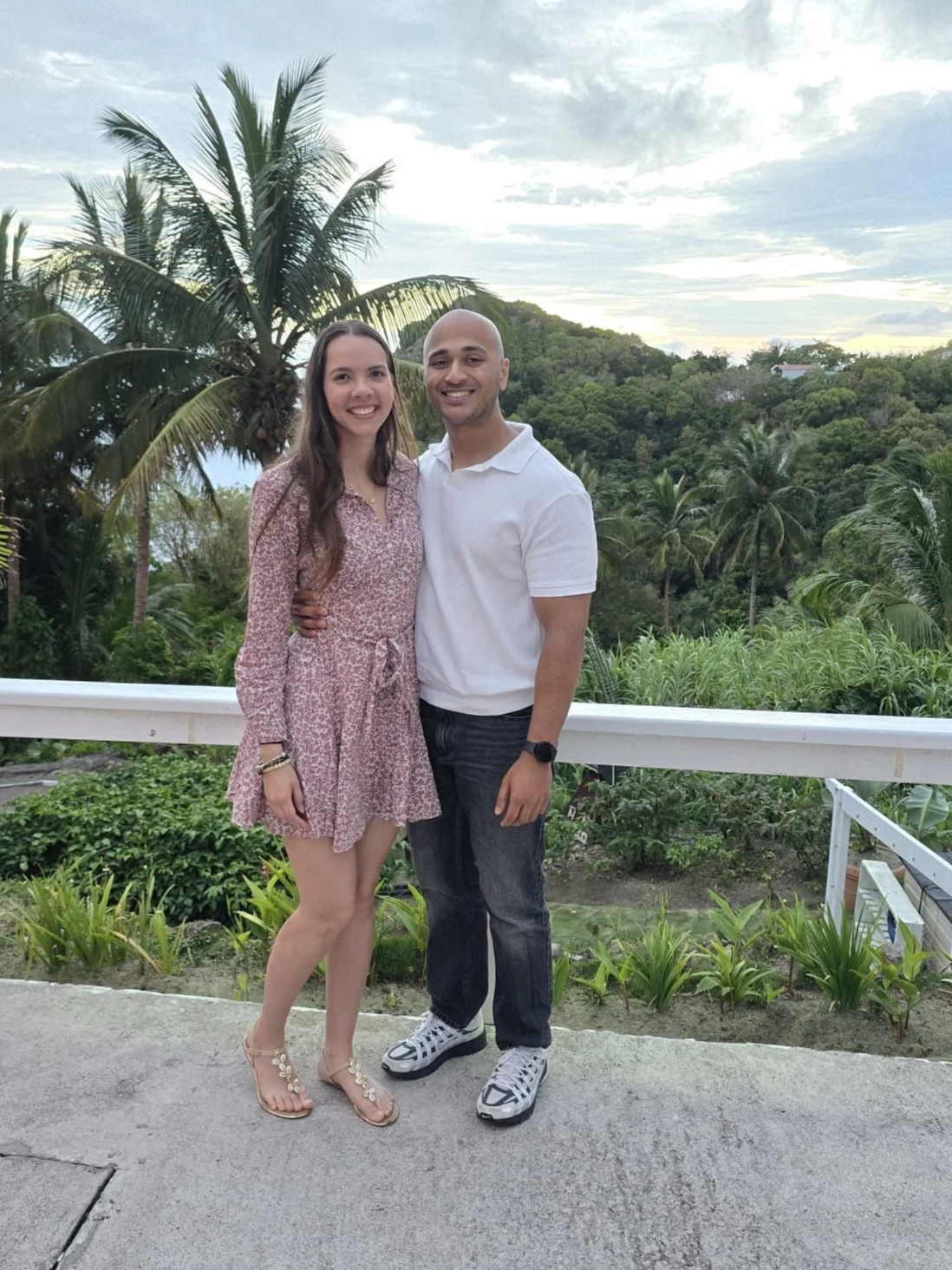 Een man en een vrouw staan samen op een terras met groene planten en palmbomen op de achtergrond, onder een bewolkte hemel.