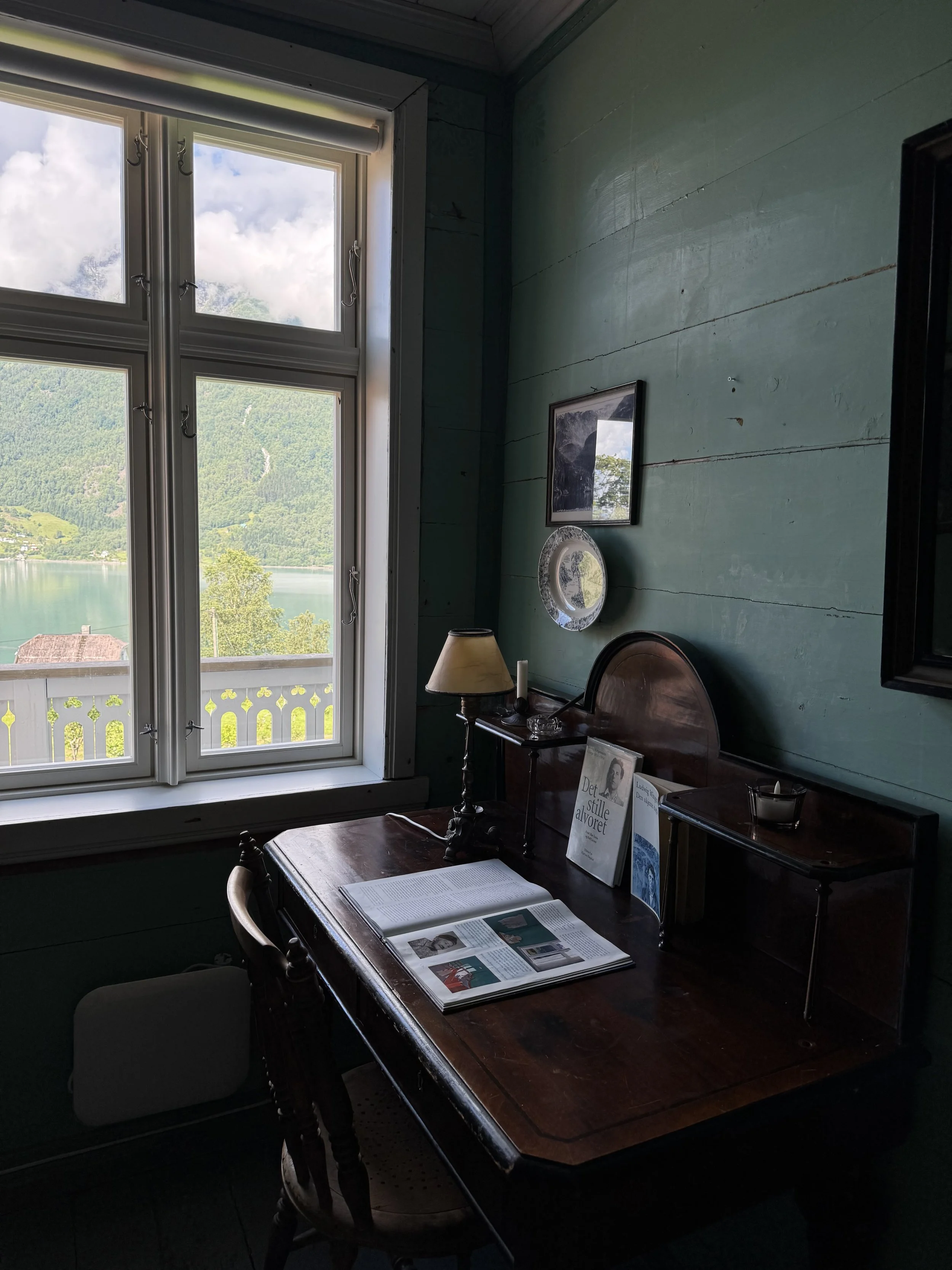 A cozy room with a wooden desk near a large window overlooking a lake and mountains. The desk has an open book, a table lamp, and some decorative items on it. The wall is painted light green with framed pictures and a decorative plate hanging on it.