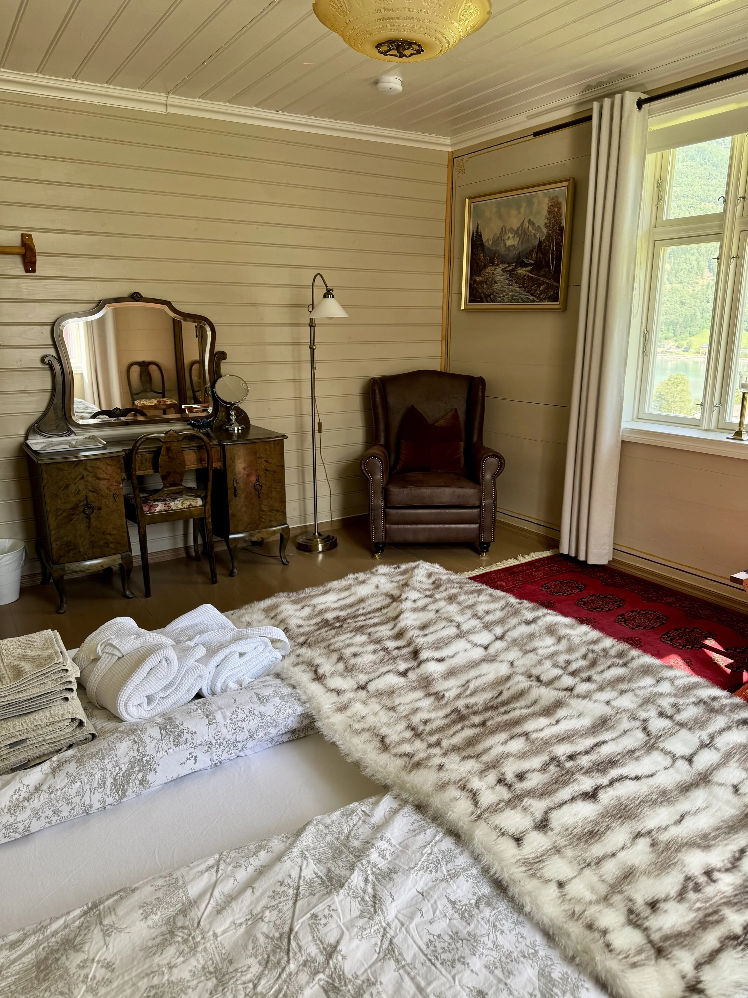 A cozy bedroom with a bed covered in a patterned duvet and a faux fur blanket. Towels are folded on the bed. There is a vintage dressing table with a mirror, a chair, and a small table with a mirror. An armchair with a pillow is positioned near a window with white curtains, and a landscape painting hangs on the wall.