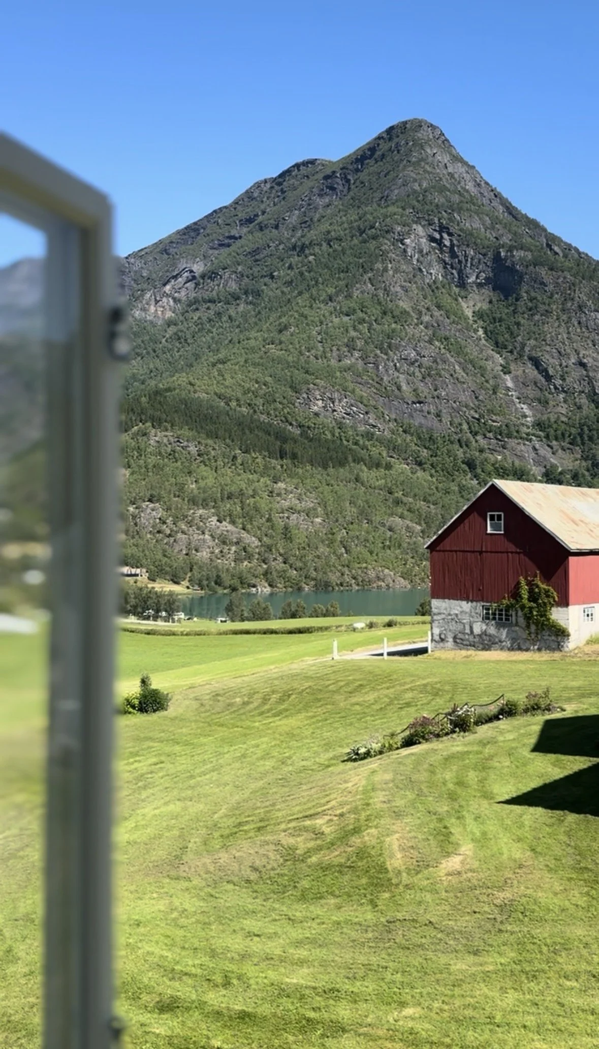 Scenic view of a red barn with a rustic roof, lush green lawn, a mountain in the background, and a body of water, seen from a window with part of the window frame visible on the left.