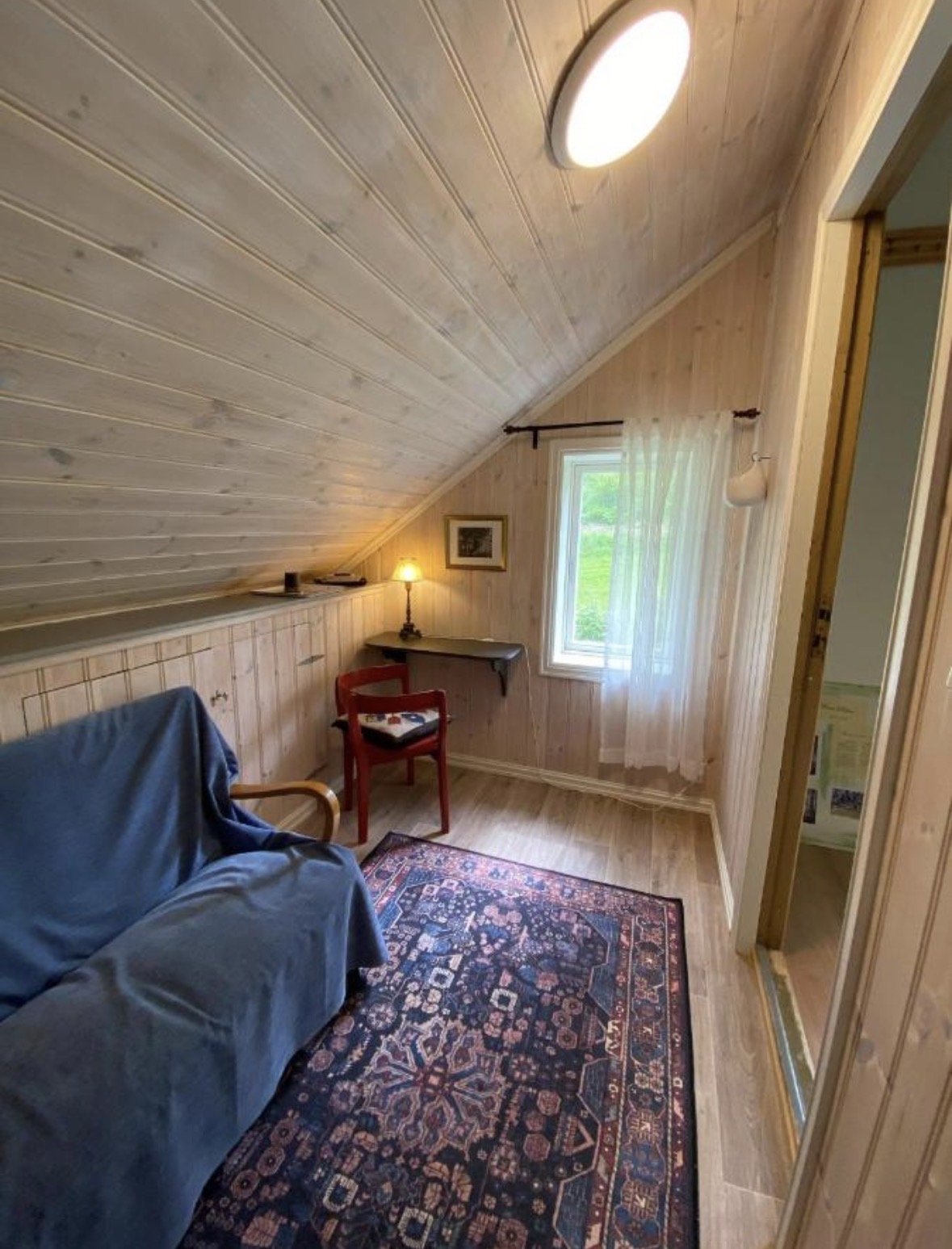 A cozy room with wooden walls and ceiling, a window with sheer curtains, a blue sofa, a red chair with a patterned cushion, a small desk with a lamp, a framed picture, a patterned rug, and a doorway leading to a bathroom.