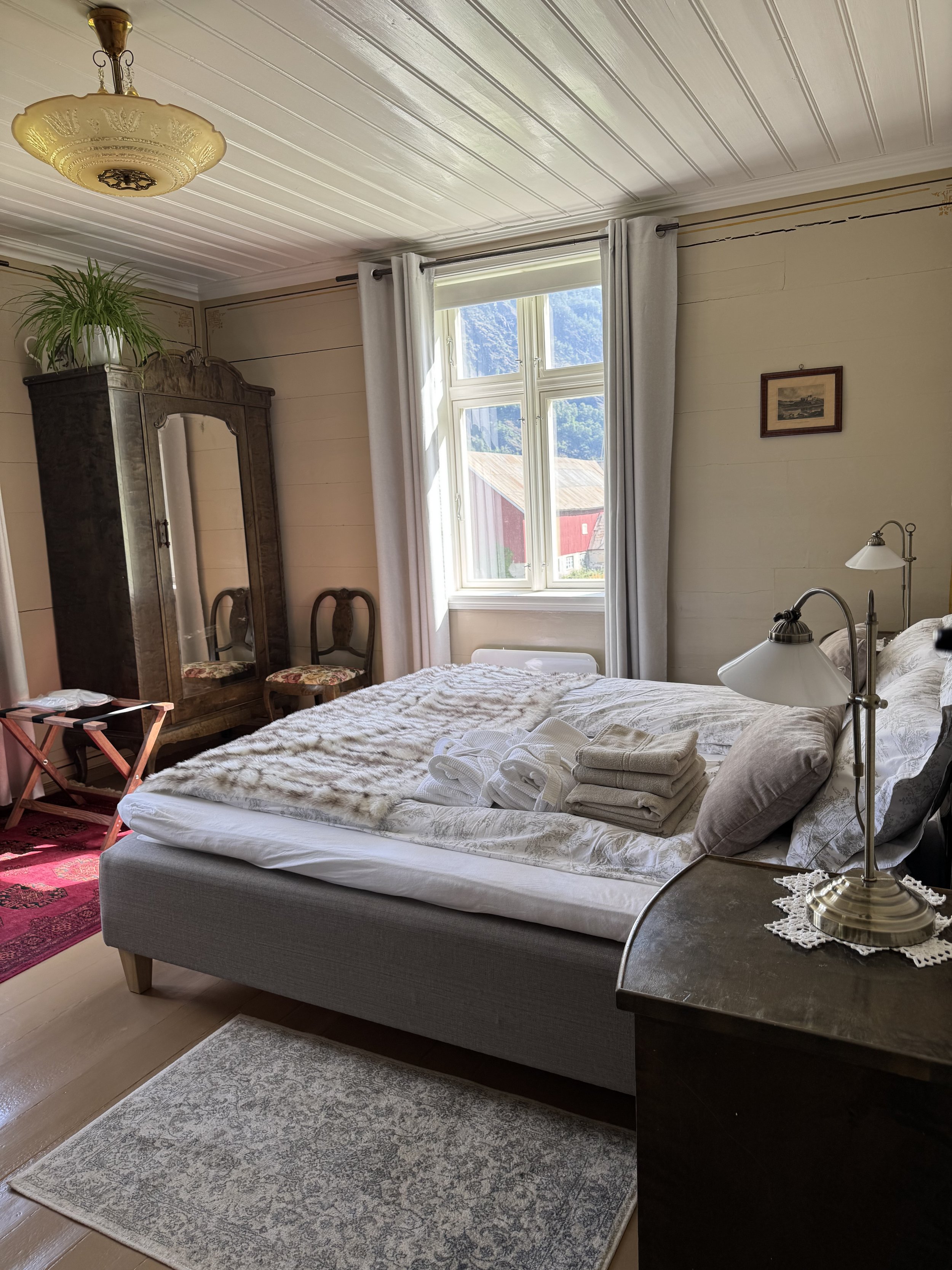 A cozy bedroom with a large bed, neatly made with white linens, pillows, and folded towels. There is a nightstand with a brass lamp and lace doily, a window with white curtains showing a mountain view, a wooden wardrobe with a mirror, and two chairs with floral upholstery. The room has a vintage style with wooden flooring, a patterned area rug, and a ceiling light fixture.