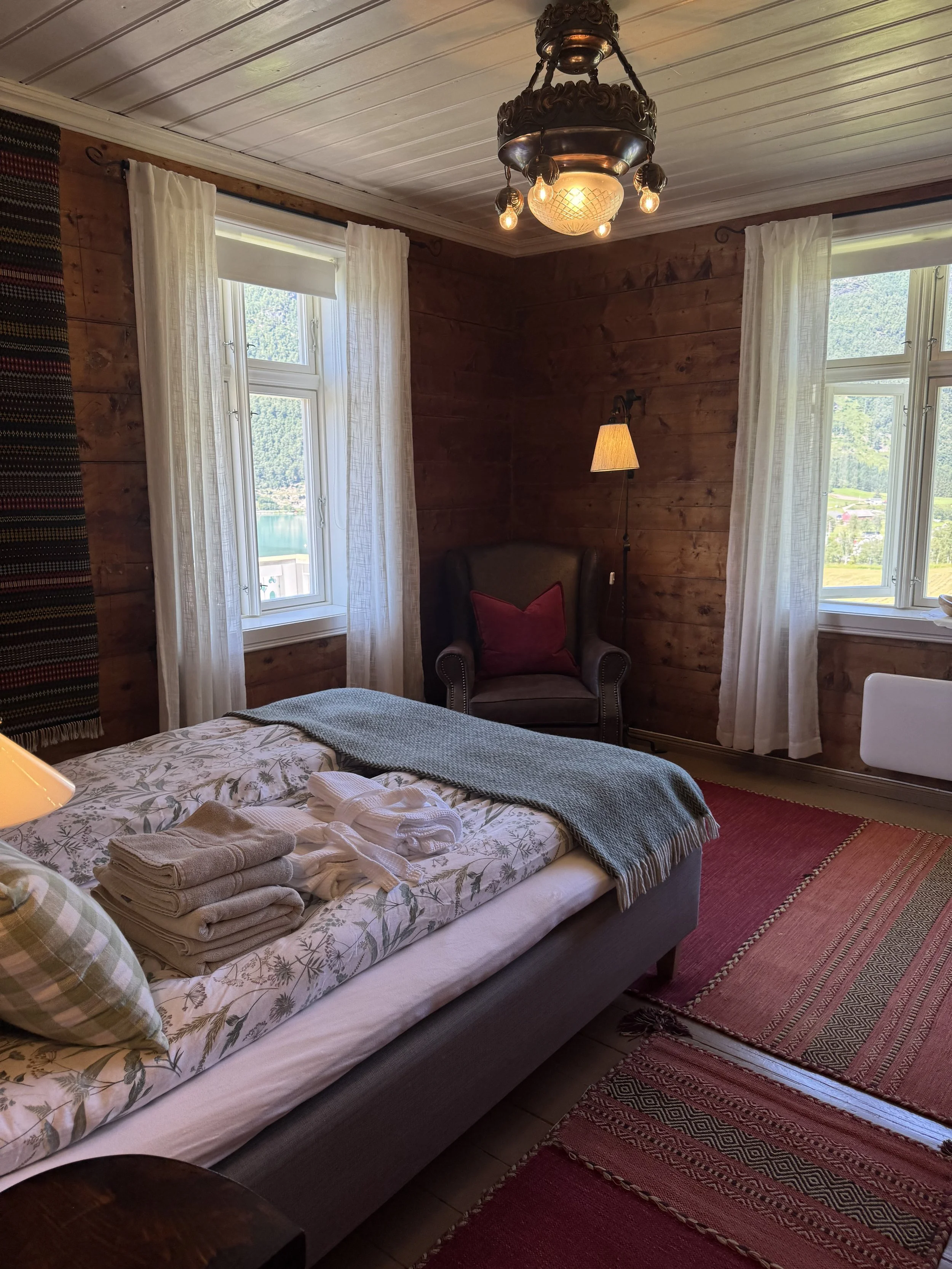 A cozy bedroom with wooden panel walls, white curtains on two windows, a bed with folded towels, a patterned pillow, and a blanket. There's a vintage chandelier hanging from the ceiling, a black armchair with a red cushion, a standing lamp, and a red and orange woven rug on the wooden floor.