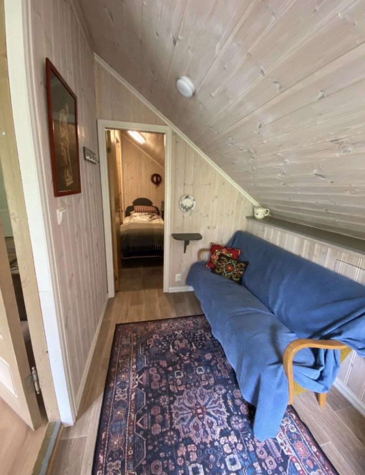 A cozy living space with wooden paneling on the walls and ceiling, a blue sofa with decorative pillows, an ornate area rug, and a doorway leading to a bedroom with a bed and wall decoration.