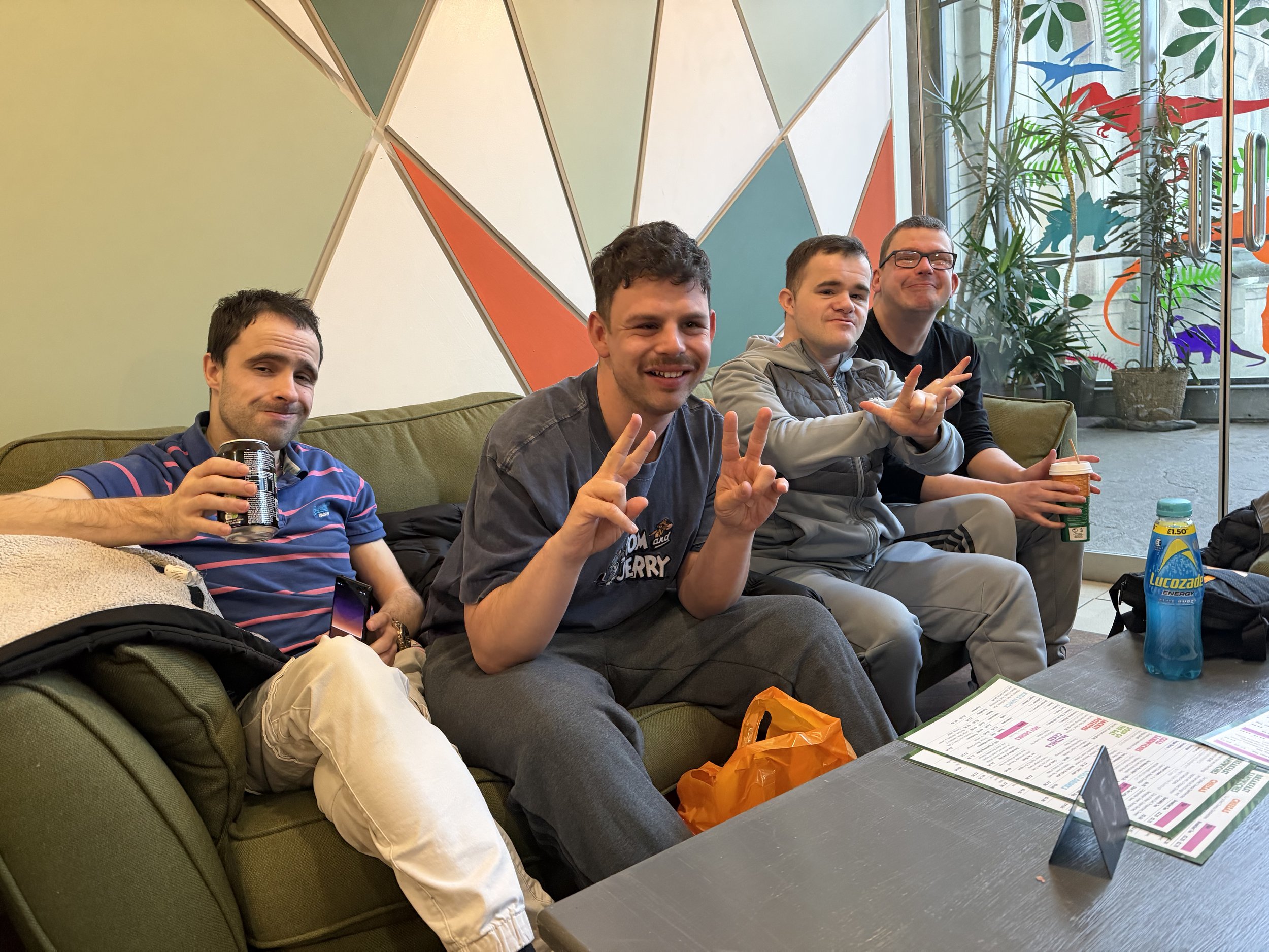 Four men sitting on a green sofa in a colorful cafe, smiling and making peace signs. The man on the far left holds a drink and a phone. the second man from the left is smiling and making a peace sign with both hands. The third man is giving a peace sign with his right hand. The fourth man holds a drink and is smiling. There are menus on the table in front, and behind them is a window with plants and colorful dinosaur decorations.
