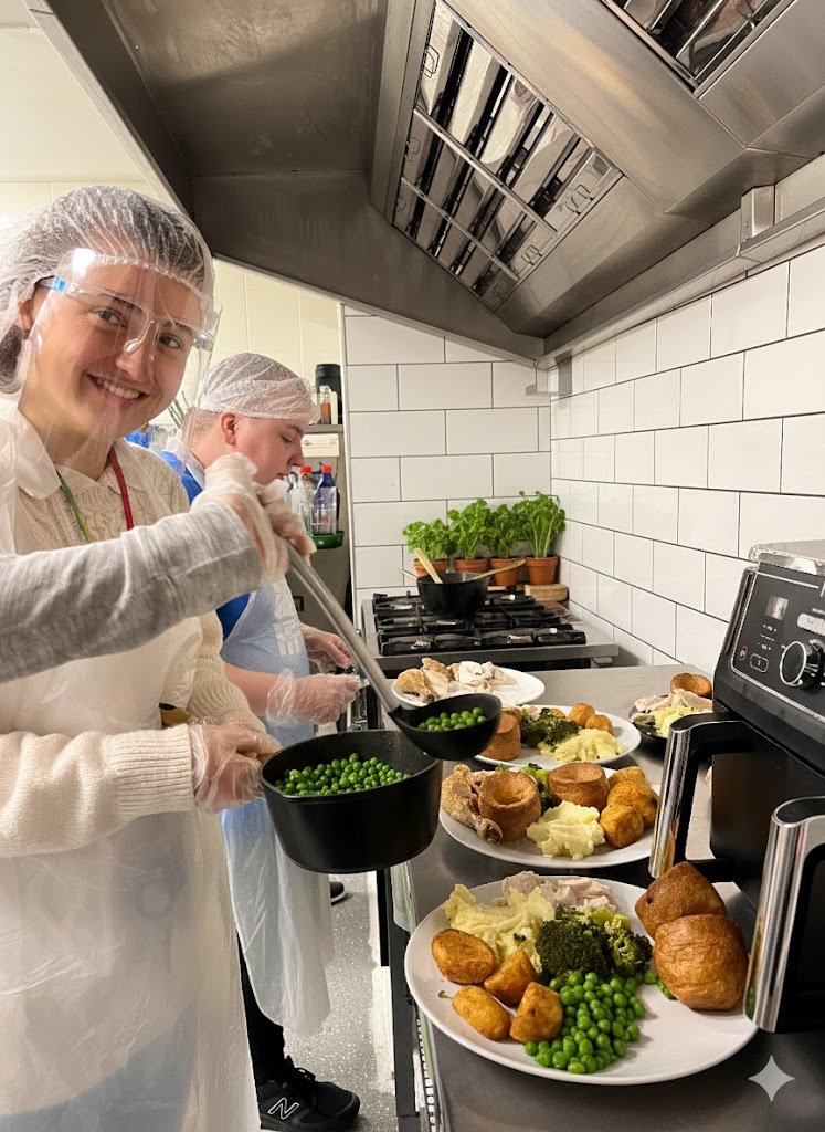 People preparing food in a commercial kitchen with plates of cooked vegetables, potatoes, and bread, wearing hairnets and gloves.
