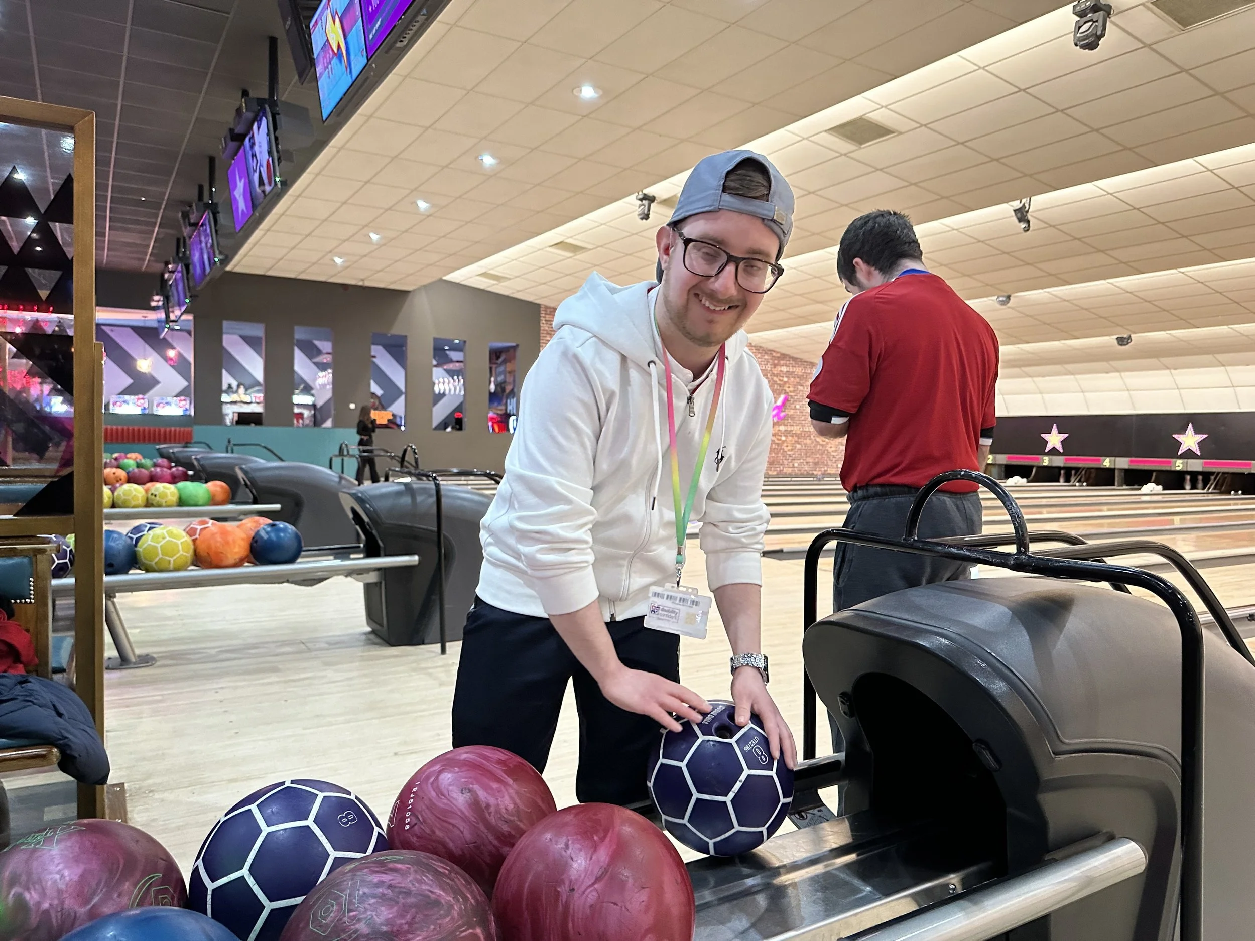 A young man in a white hoodie, wearing glasses and a backwards cap, at a bowling alley, holding a purple bowling ball, smiling.
