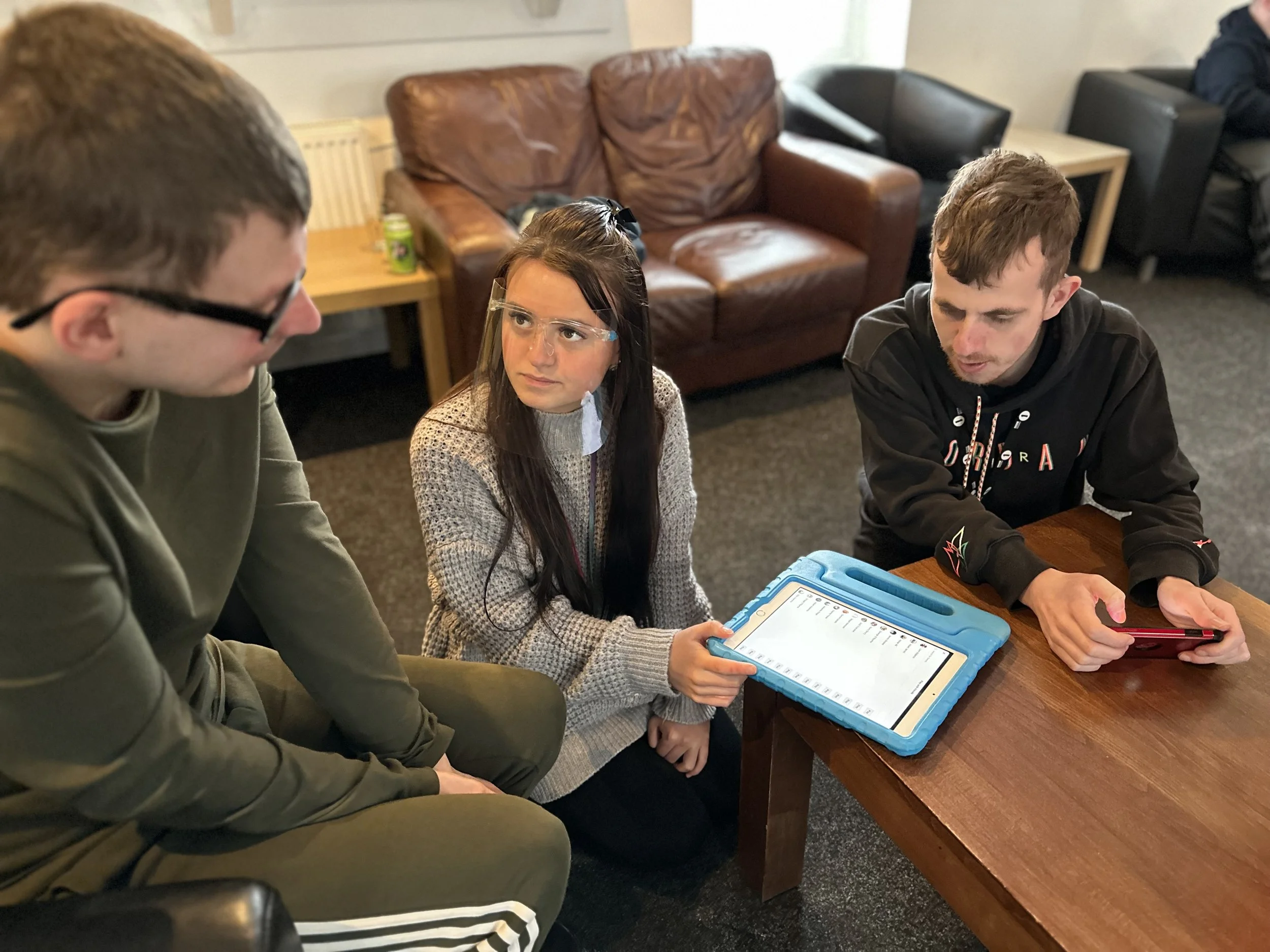 A group of four people gather around a wooden table indoors, with a woman sitting on her knees, holding a tablet, and three men, one with glasses leaning forward, all engaging in a conversation.