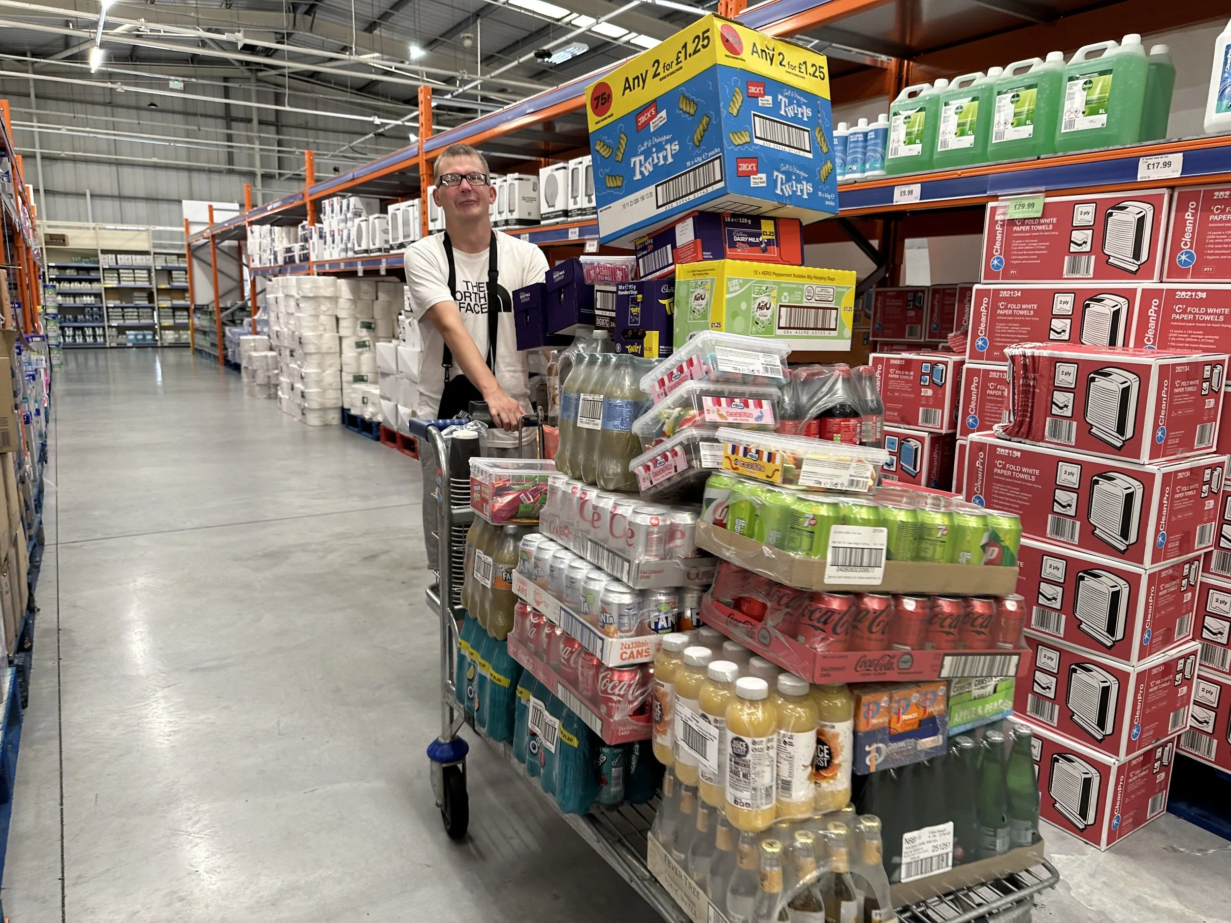 A young man wearing glasses and a white t-shirt shopping at a wholesale store, upright on the aisle with a cart filled with beverages, snacks, and bottled water, surrounded by shelves stocked with grocery items.