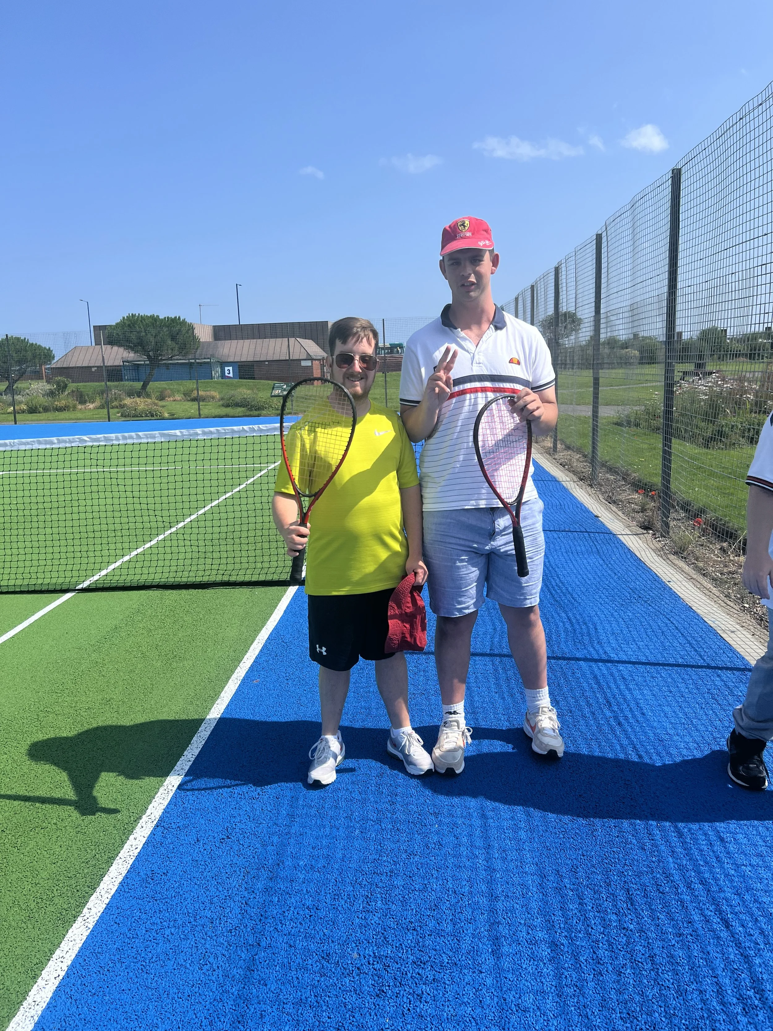 Two males standing on a tennis court, each holding a tennis racket, with a clear blue sky above. One is wearing a bright yellow shirt and sunglasses, the other is wearing a white striped shirt and a red cap, both smiling.