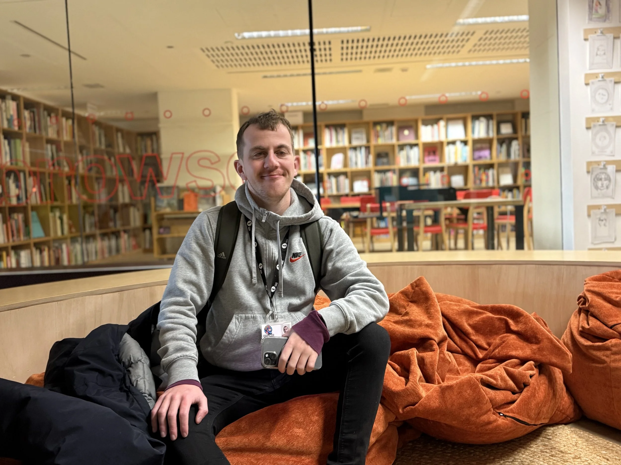 A young man sitting on an orange bean bag chair inside a library, smiling and holding a smartphone, with bookshelves and tables in the background.