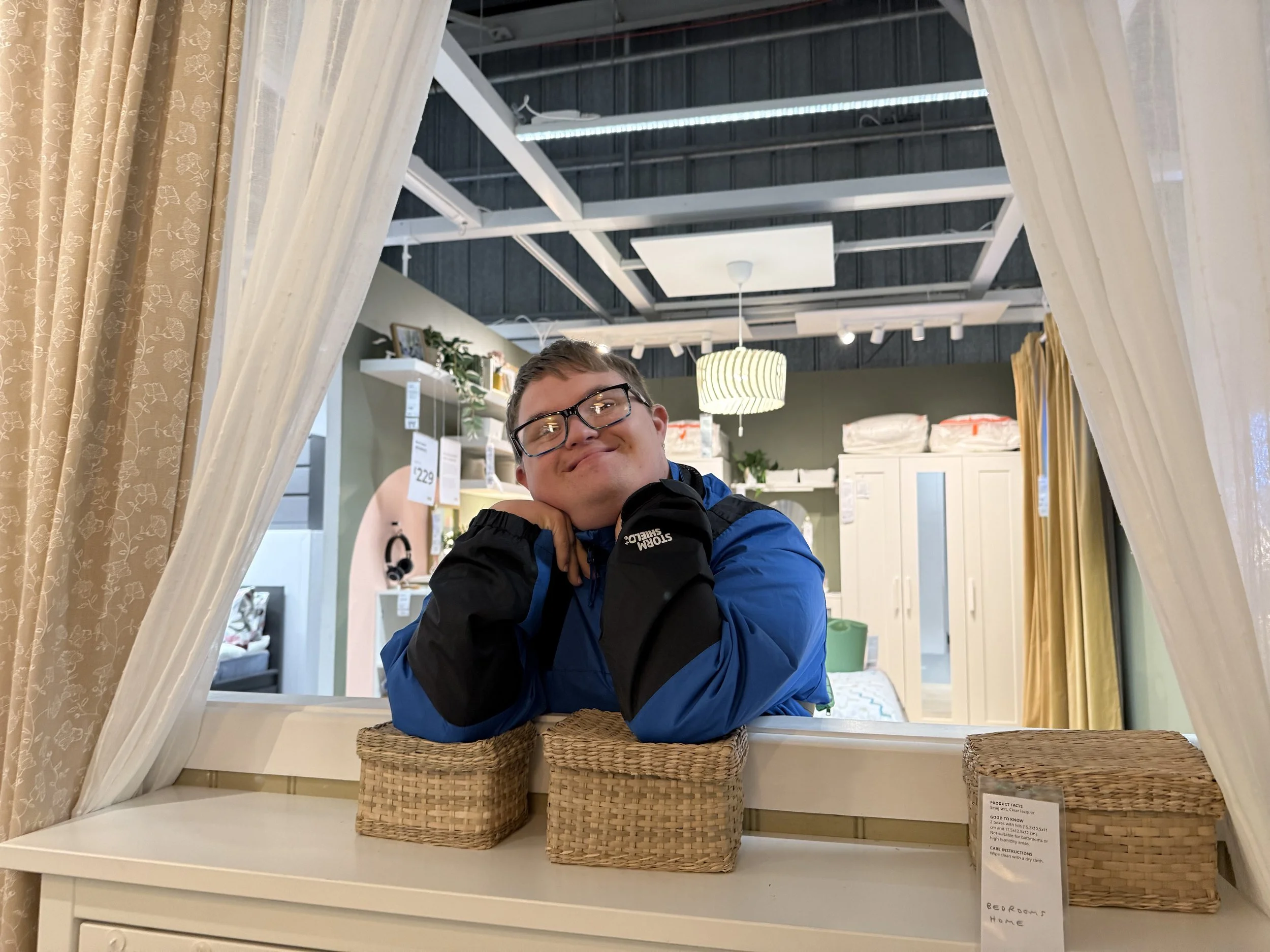 A person with glasses and a blue jacket is resting their chin on their hands, leaning on a white piece of furniture, and smiling at the camera. The scene is inside a store or showroom with furniture and home decor items visible in the background, framed by curtains and a mirror.