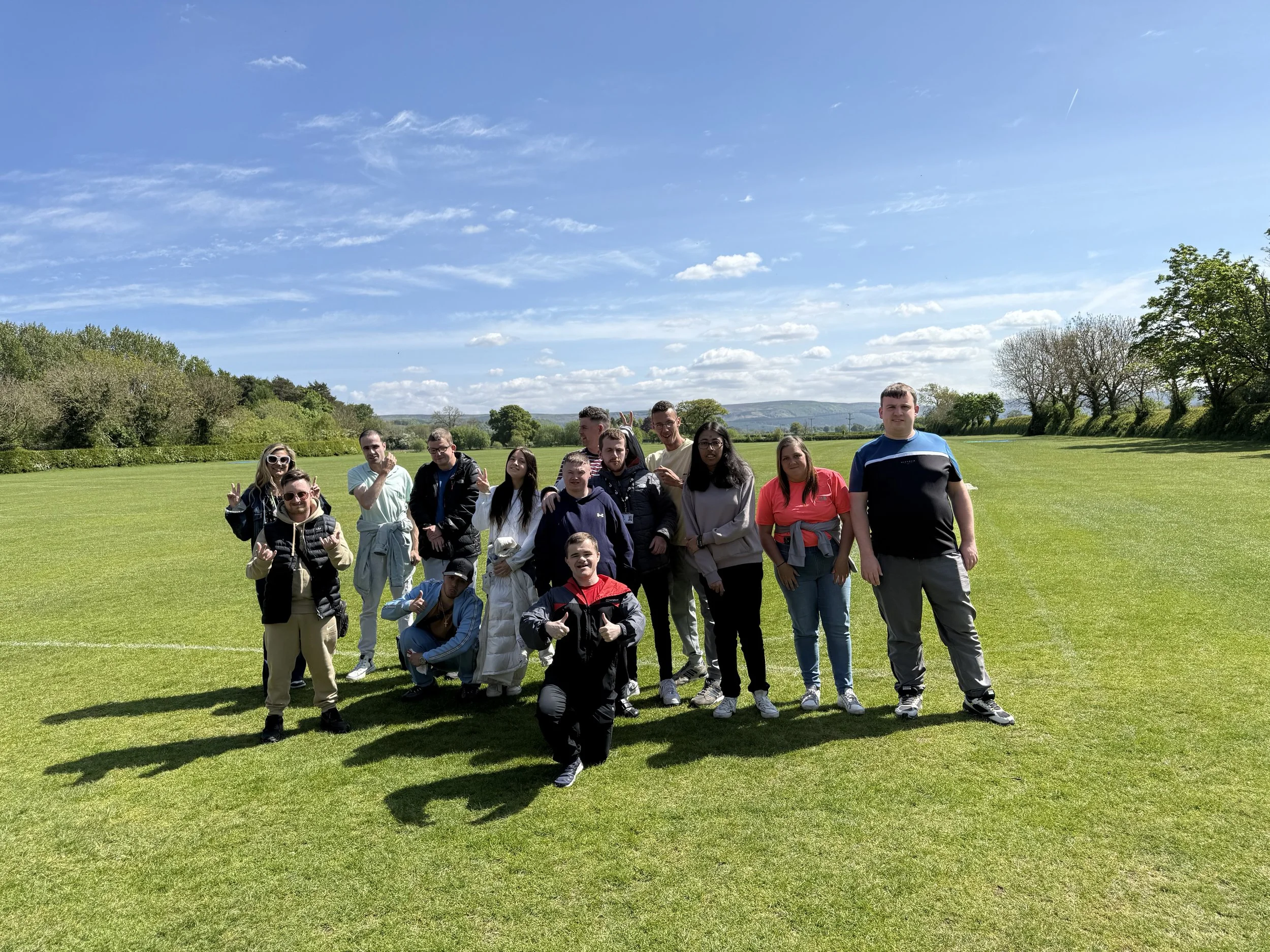 Group of people outdoors on a sunny day standing on a grassy field with trees in the background.