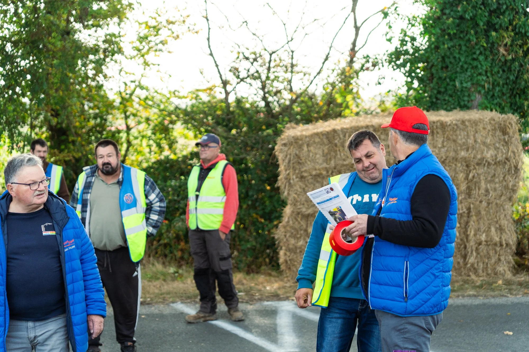 Groupe de personnes portant des gilets de sécurité, conversations en plein air près de foin empilé, arbres en arrière-plan.