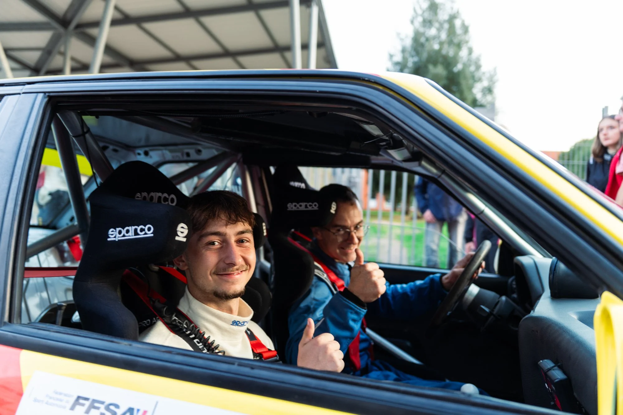 Deux hommes dans une voiture de course, les ceintures de sécurité, l'un fait un signe d'appréciation et le second donne un pouce en l'air, avec un groupe de spectateurs en arrière-plan.