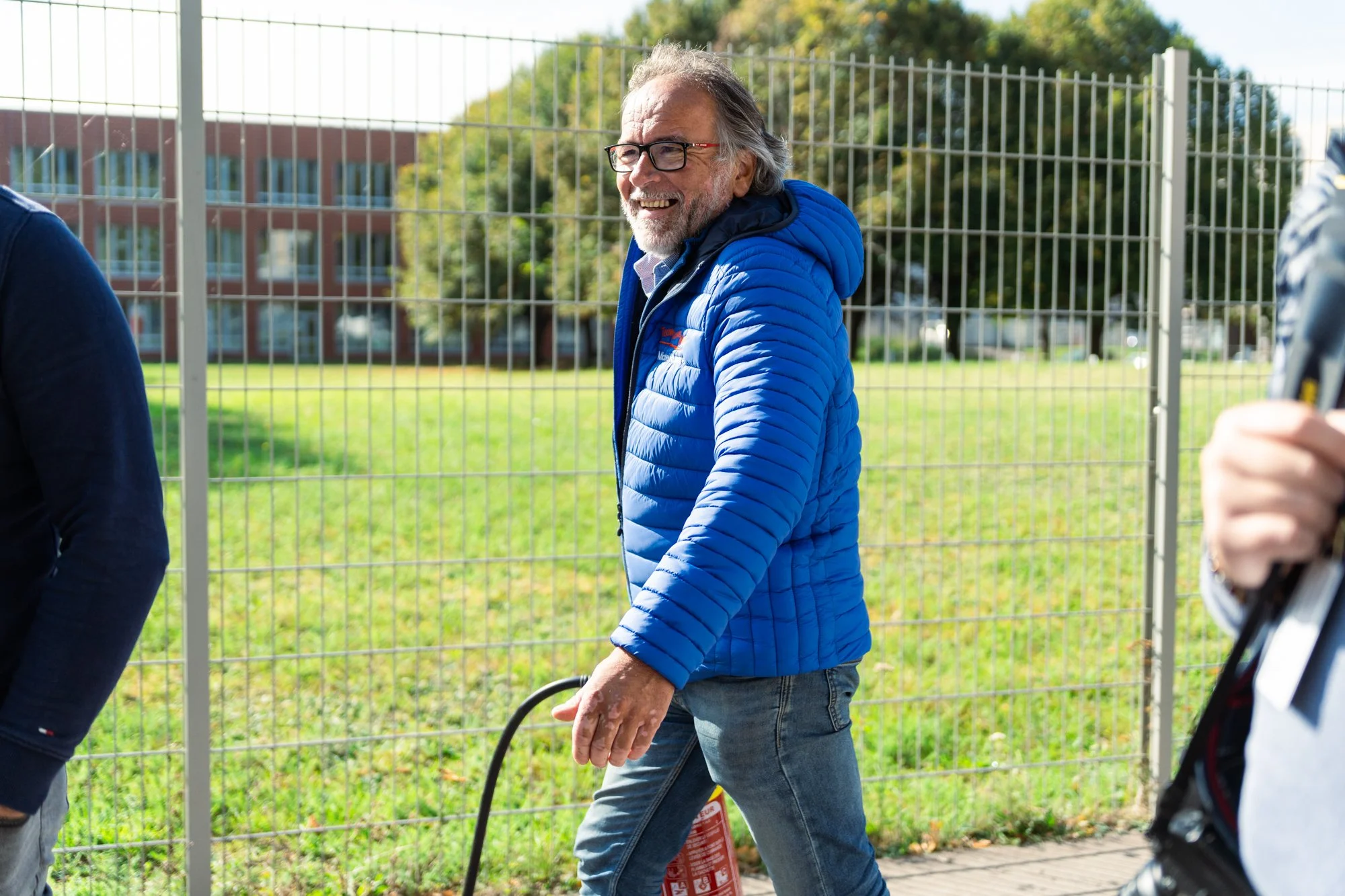 Un homme âgé avec des cheveux longs, une barbe grise, portant des lunettes noires, un manteau en duvet bleu, marche à côté d'une clôture en métal, lors d'une journée ensoleillée dans un parc.