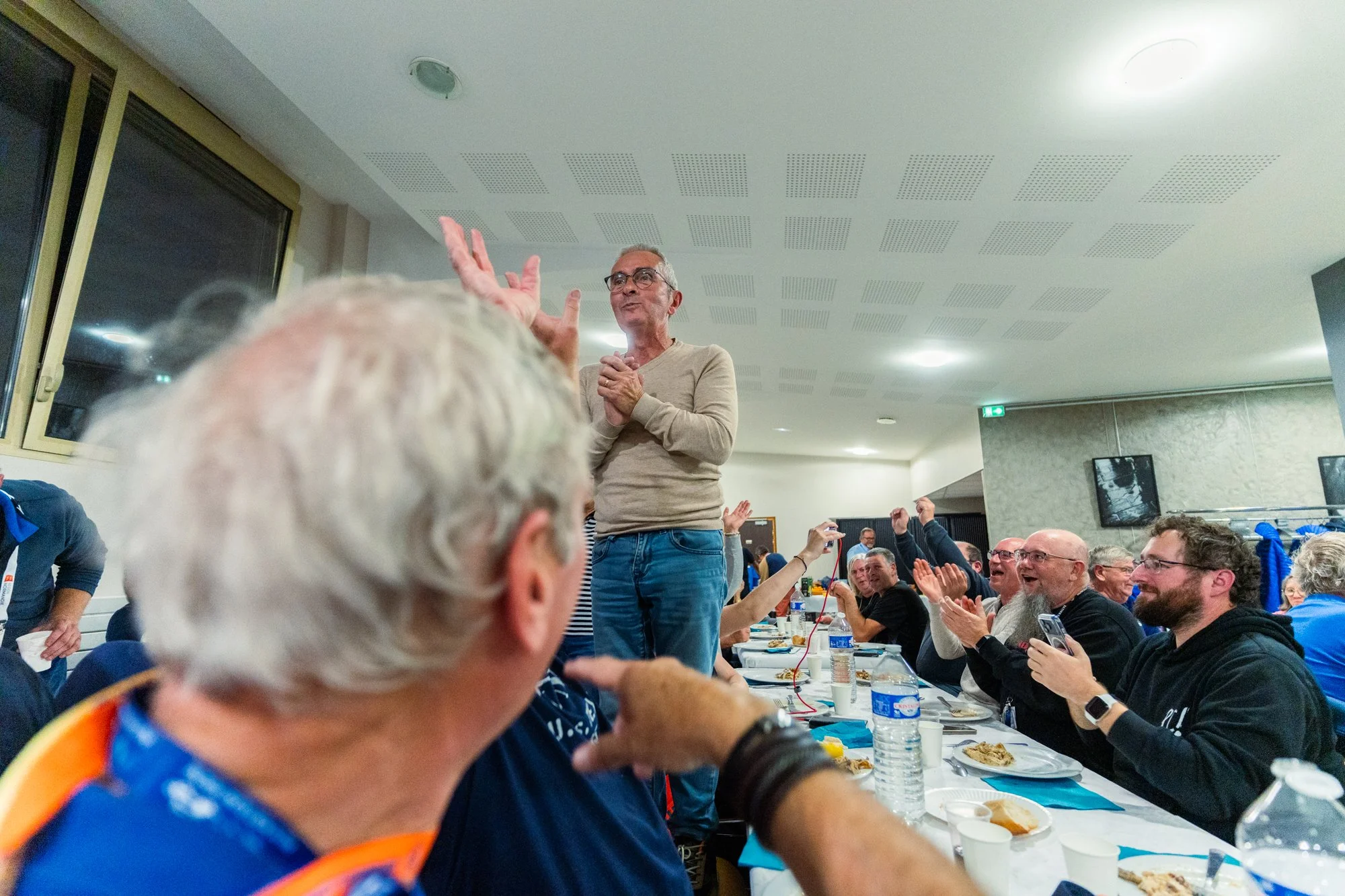 Une réunion ou un rassemblement de personnes dans une salle, avec un homme debout en train de parler ou de s'adresser à l'audience, entouré de personnes assises à une table, applaudissant, avec des plats et des bouteilles d'eau sur la table.