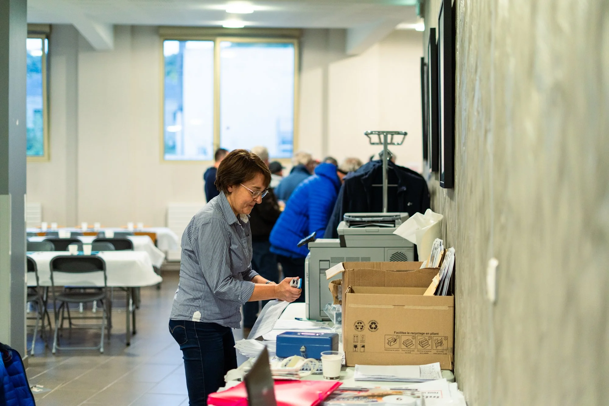 Une salle avec des personnes en train d'imprimer ou de préparer des documents. Une femme en premier plan tient un téléphone ou un dispositif portable, devant une caisse enregistreuse et des cartons remplis de papiers.