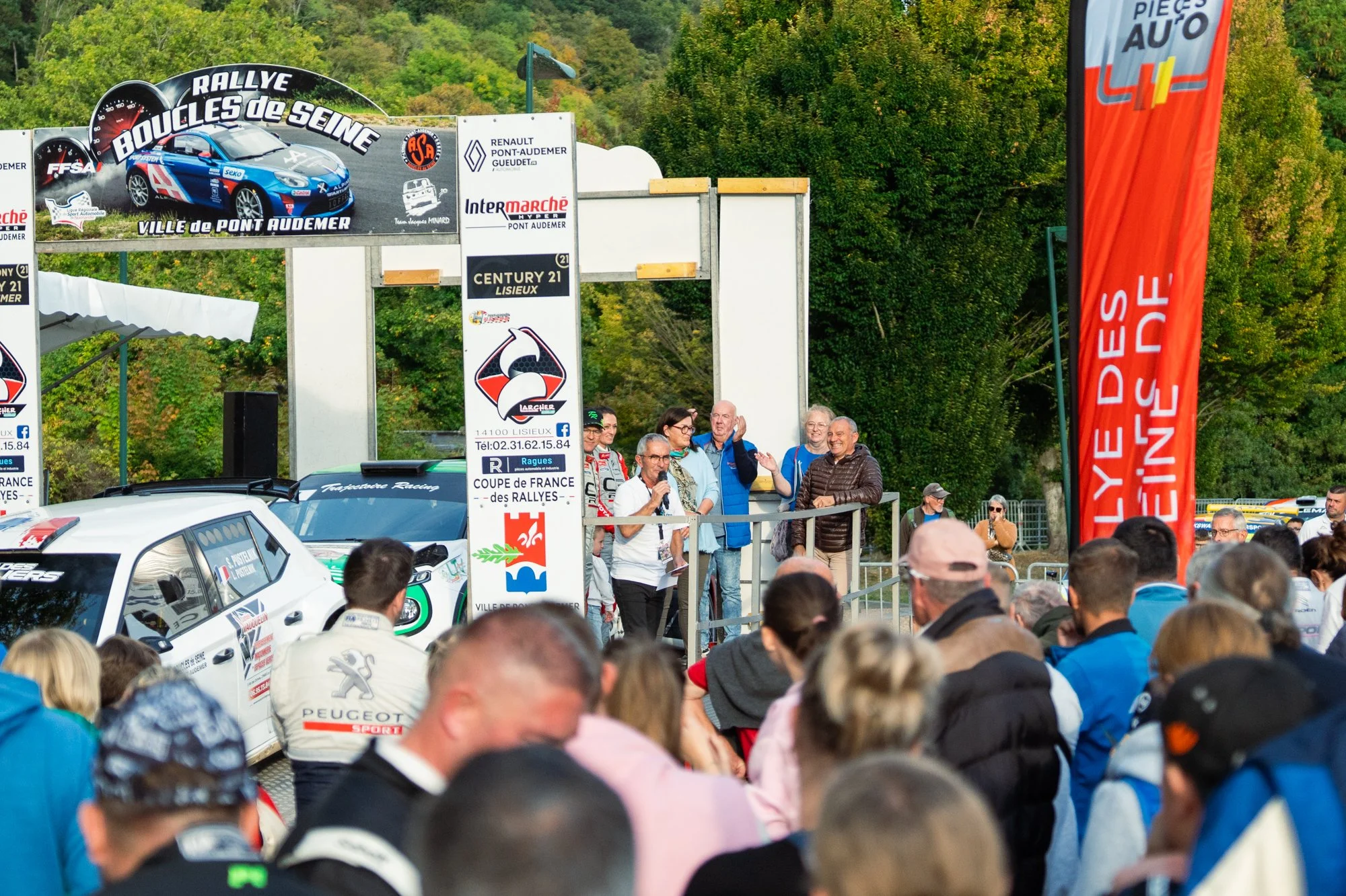 Une scène d'un rallye automobile avec des spectateurs et des voitures de course, un panneau publicitaire, et des personnes parlant ou étant interviewées. Il y a des arbres en arrière-plan et une grande bannière rouge à droite.