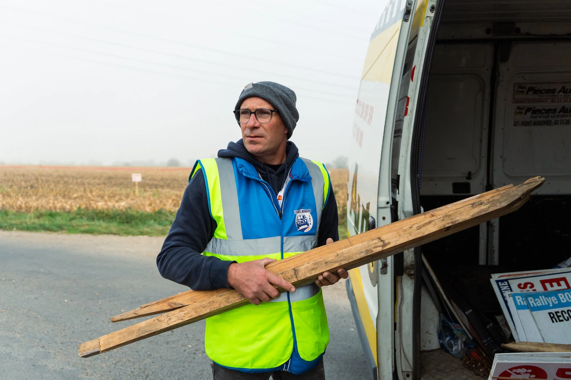 Un homme portant un gilet de sécurité jaune et un bonnet gris se tient à côté d'un camion de livraisons, tenant une planche de bois dans ses mains.