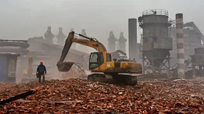 Construction site with excavator and worker, industrial buildings in background.