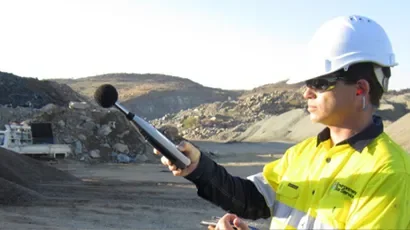 A construction worker wearing a yellow safety jacket, white hard hat, and sunglasses, holding a sound level meter in an outdoor construction site with rocky terrain and hills in the background.