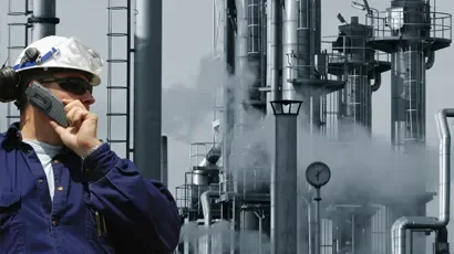 A worker in safety gear using a walkie-talkie at an industrial plant with smokestacks and pipes emitting smoke or steam in the background.