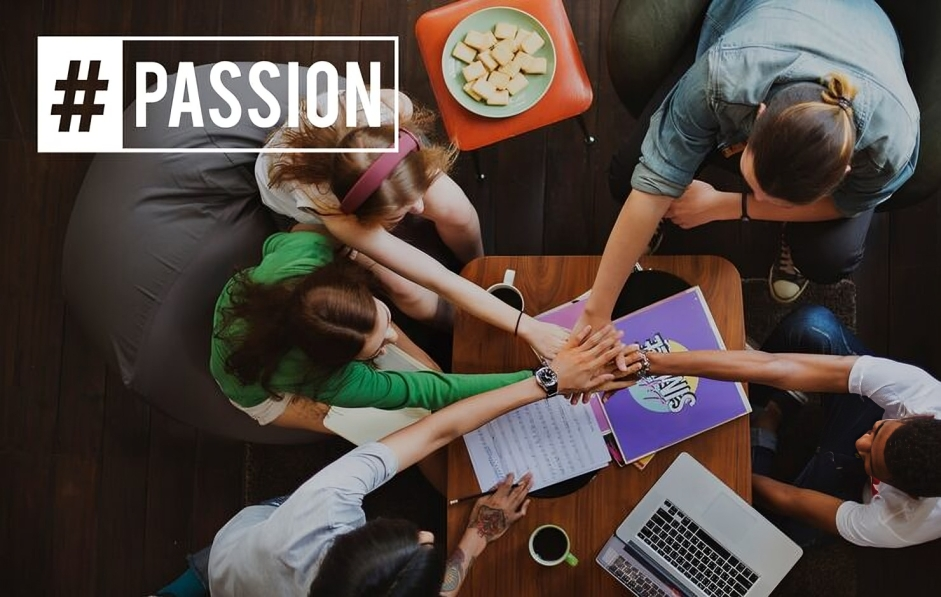 A group of people gathered around a table, joining hands in the middle, with documents, a laptop, a coffee cup, and a plate of snacks visible on the table, symbolizing teamwork and collaboration.