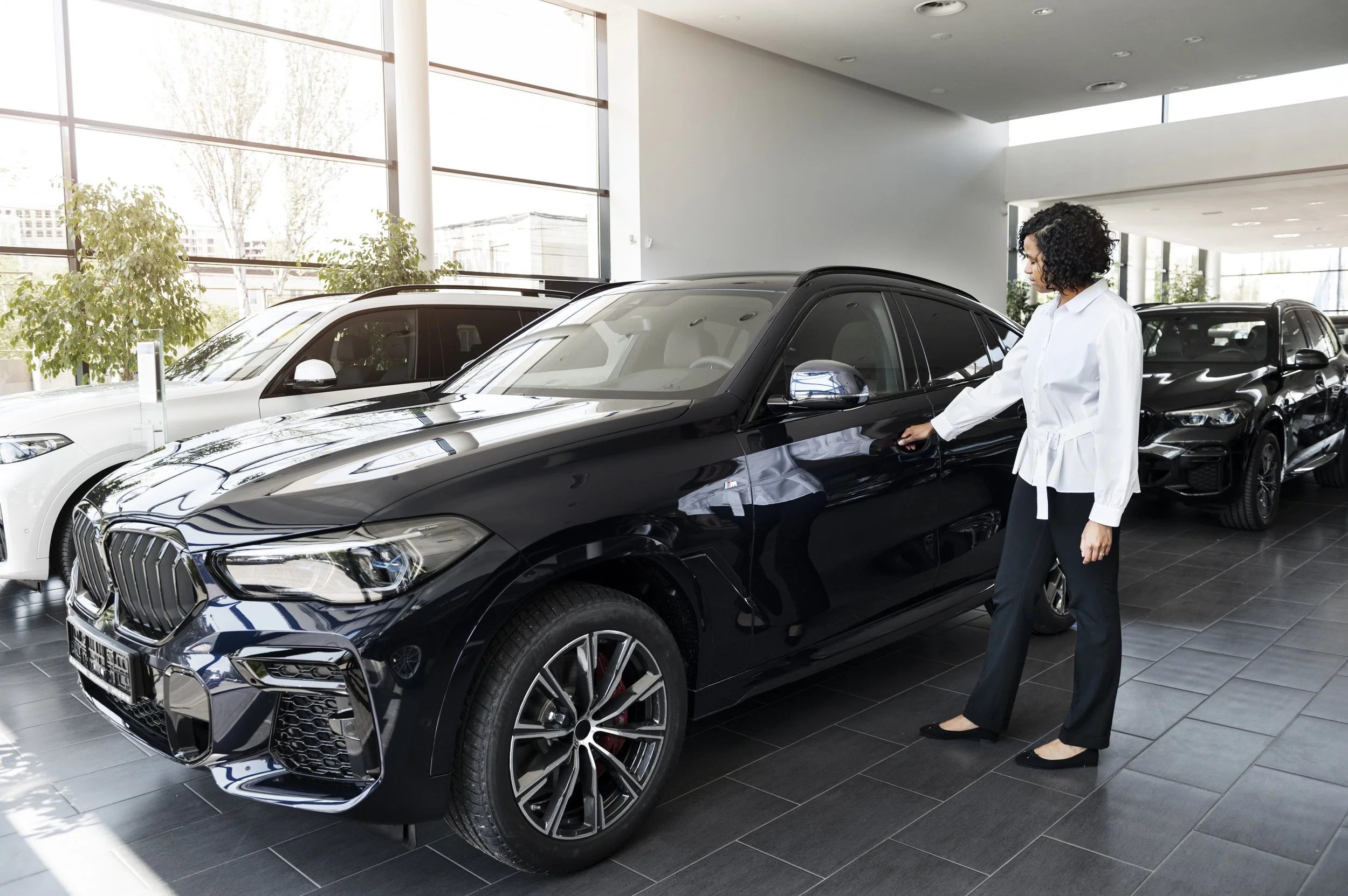 A woman in a white blouse and black pants shopping for a black BMW in a car dealership.