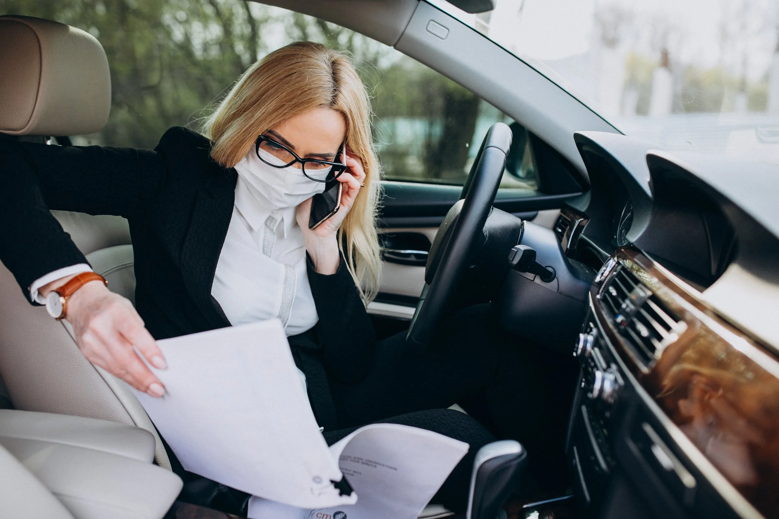A woman with blonde hair, glasses, and a face mask, dressed in a black blazer and white shirt, talking on her cellphone while sitting in the driver's seat of a car. She is holding papers and appears to be working or discussing something related to documents.
