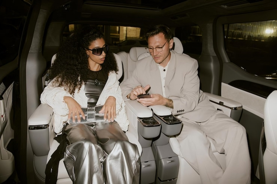 Two people sit in the back of a luxurious vehicle, looking at a smartphone together. The woman is wearing sunglasses and metallic pants, and the man is dressed in a light-colored suit and glasses.