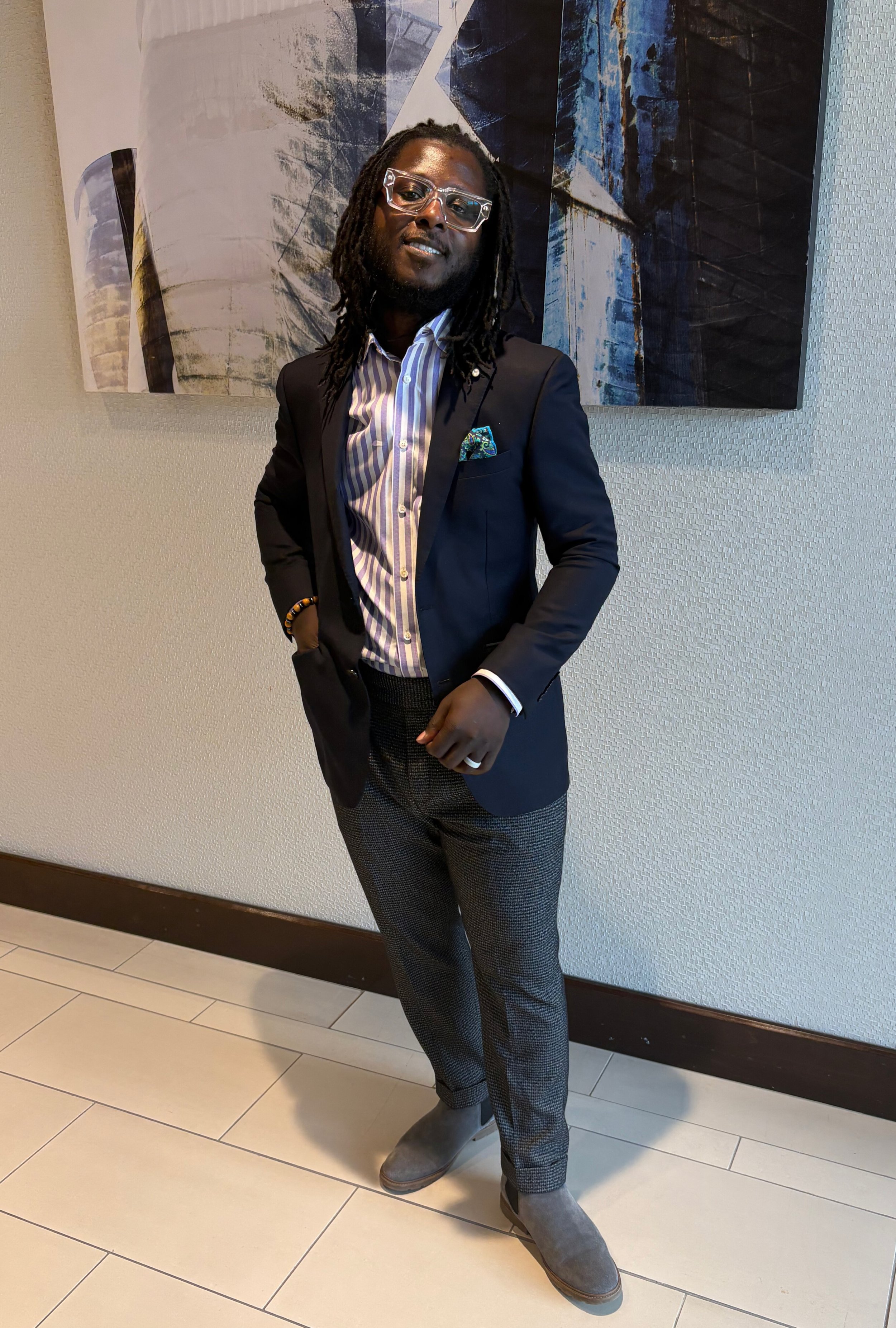 A man with dreadlocks and glasses posing in front of an abstract painting. He is dressed in a black blazer, striped shirt, patterned pants, and gray suede shoes, with one hand in his pocket and the other resting on his stomach.