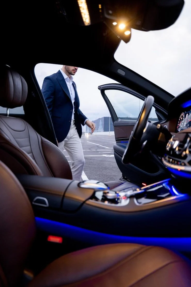 A man in a blue blazer and beige pants standing outside a luxury car parked in an open lot, viewed from inside the car with brown leather seats and illuminated blue interior lights, overcast sky in the background.