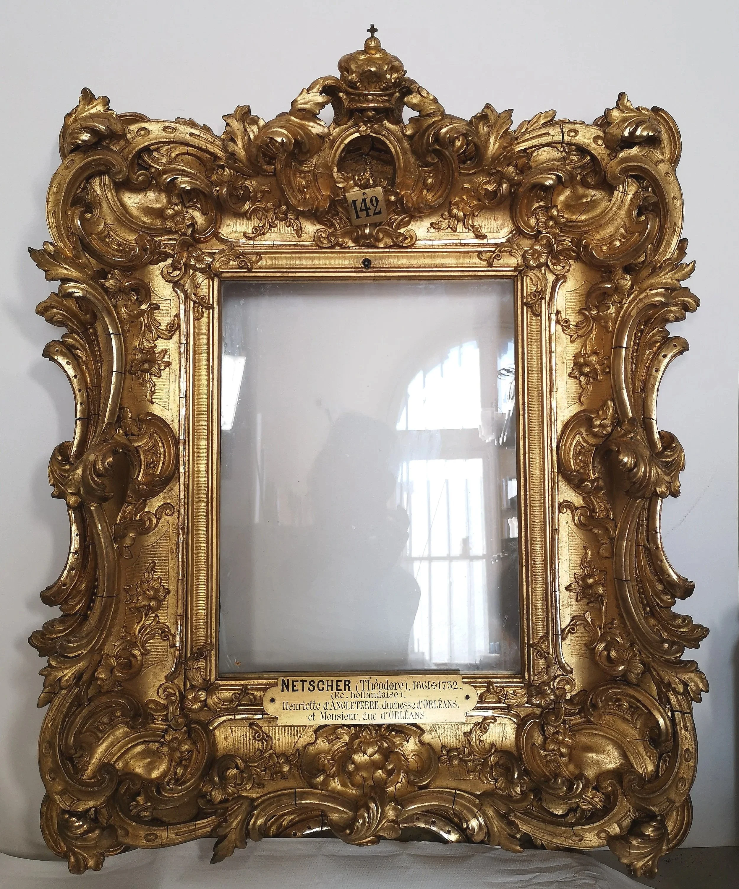 Un cadre de miroir orné de détails rocaille et ornementaux, avec une plaque en bas indiquant le nom et la date de l'artiste, ainsi que plusieurs notes à main levée. Le cadre est en bois doré très détaillé.