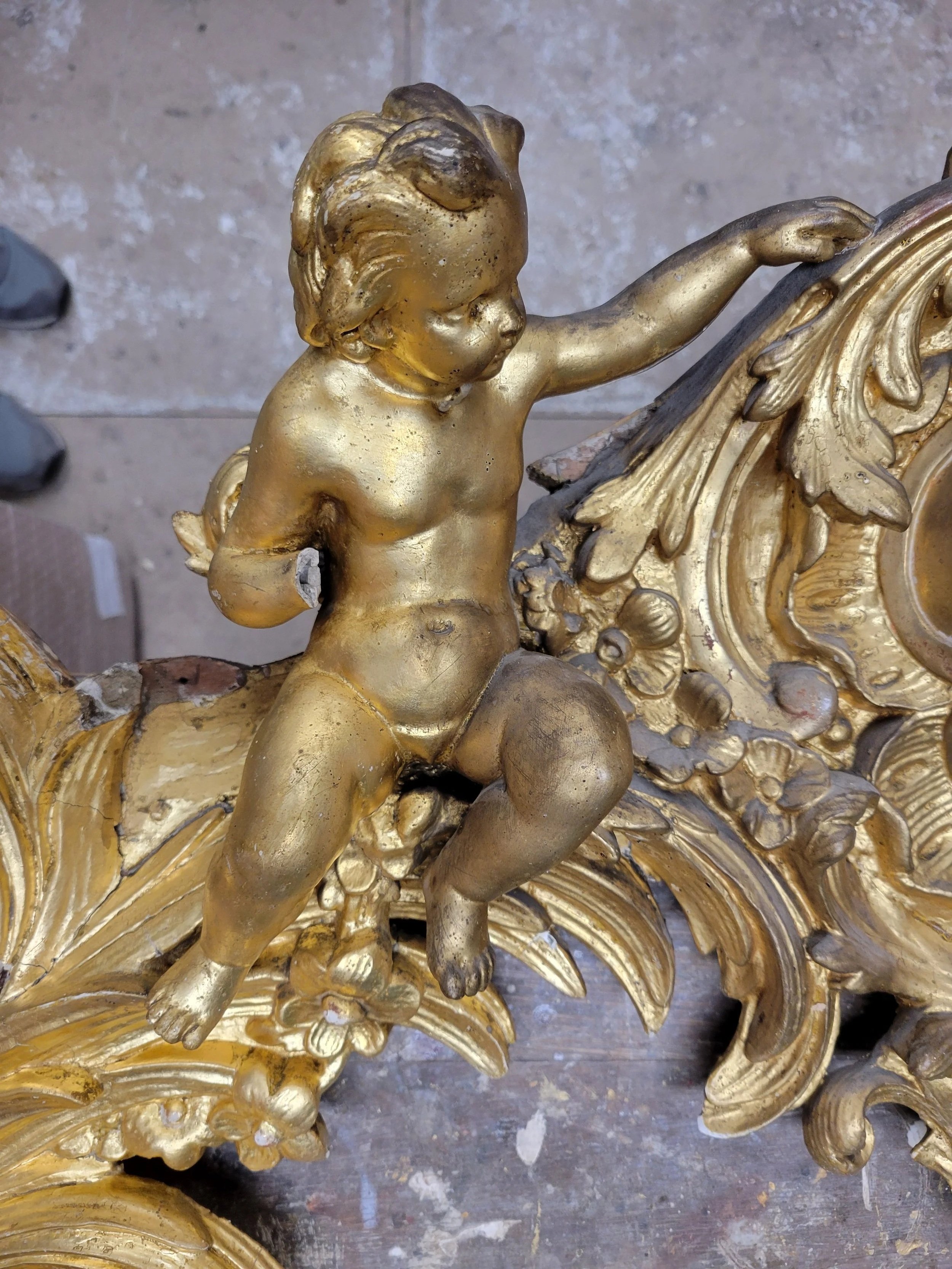 Statue en or représentant un enfant avec des ailes, assis sur une décoration ornementale.